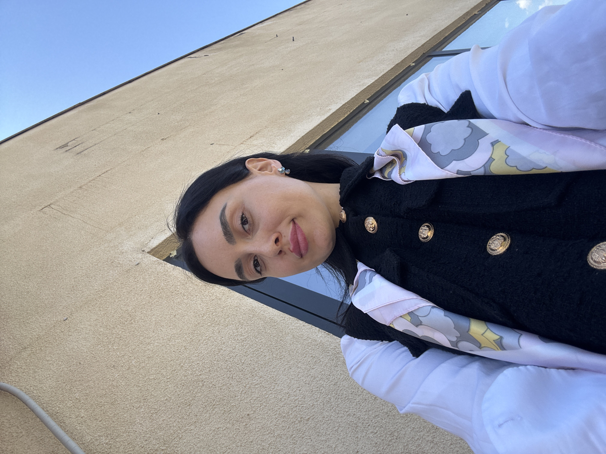 The image, oriented vertically, captures a close-up of a woman outdoors on a bright day in Tbilisi, Georgia. She is positioned in the lower left portion of the frame, looking directly towards the camera from an upward angle.
The woman has dark hair, styled down, and dark, well-defined eyebrows. Her lips are painted a soft pink, and a small, shiny stud earring is visible on her right ear. She is wearing a black, textured jacket or coat, possibly tweed or bouclé, which is adorned with several large, prominent round gold buttons. These buttons feature an intricate embossed design, possibly a lion's head or a crest, though specific details are unclear. Underneath her jacket, a crisp white long-sleeved shirt is visible. A patterned silk scarf, featuring abstract or cloud-like shapes in shades of pink, grey, and yellow, is stylishly tucked into her neckline.
The background is dominated by a light beige or tan stucco-like building wall, which appears textured and shows some minor marks. A narrow dark stripe, possibly a window frame or structural element, runs vertically behind her, separating the wall from a clear blue sky that occupies the upper right corner of the frame. In the far bottom left corner, a grey ribbed hose or conduit is visible against the wall.
The activity is a static pose for a photo. The time of day appears to be daytime, given the clear blue sky and bright, even lighting, suggesting sunny weather. The woman's expression is calm and neutral, with a slight hint of a smile. There is no visible text in the image.