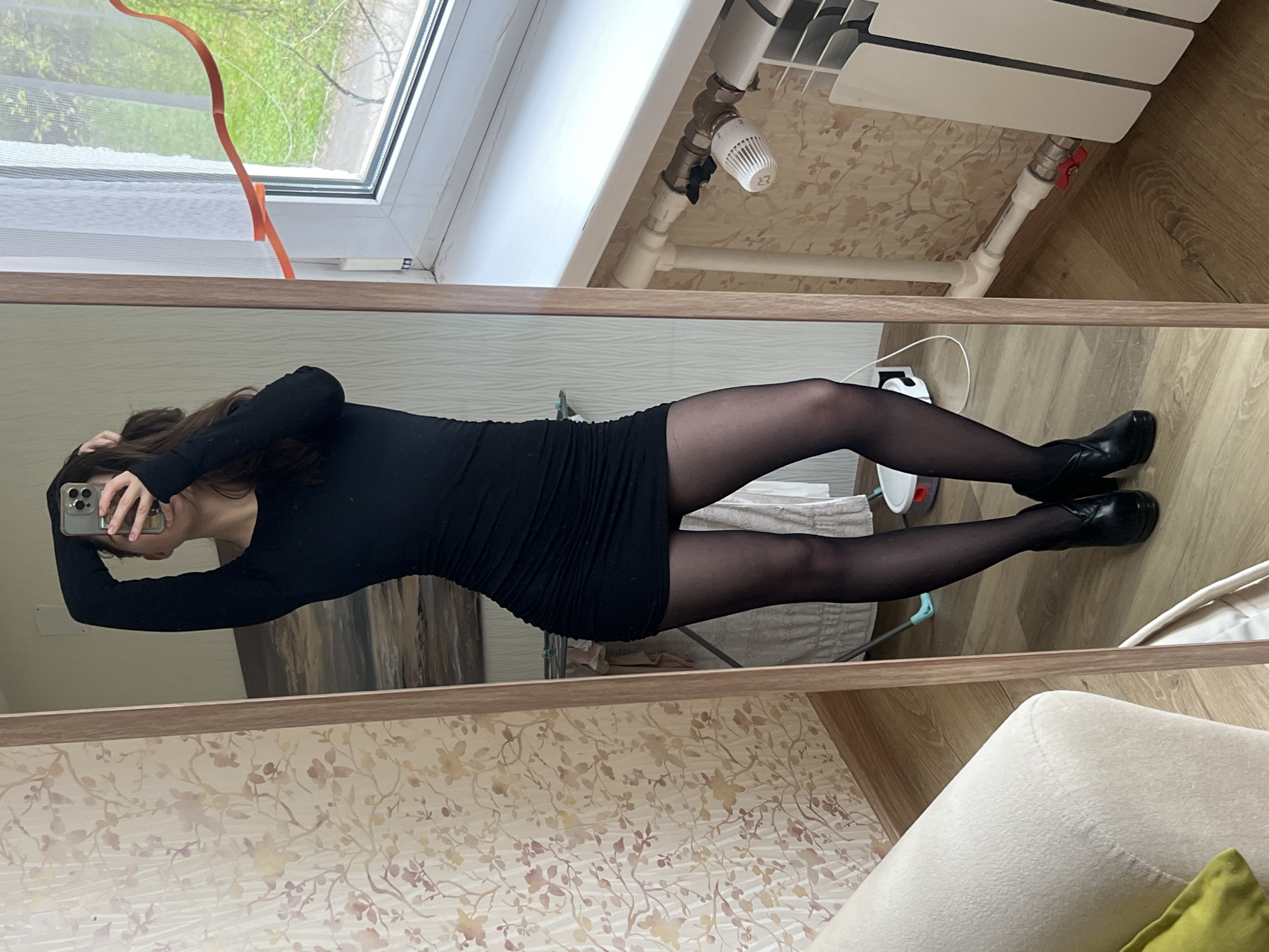 The image displays a mirror selfie of a woman taken indoors. The woman, with long dark hair, is seen holding a silver-colored smartphone with multiple camera lenses, obscuring part of her face as she captures her reflection. She is wearing a fitted black long-sleeved mini-dress, sheer black pantyhose or tights, and black shoes, possibly low-heeled boots.
The reflection in the mirror reveals various elements of the room. Behind the woman, a window offers a view of green foliage and what appears to be a tree outside, suggesting daytime. An orange ribbon or piece of tape is visible near the top of the window frame. Below the window, a white radiator with exposed pipes and a thermostatic valve is mounted against a light-colored wallpaper adorned with a subtle floral or branch pattern. The number "23" is visible on the thermostat. The flooring is light-colored wood laminate. In the bottom right corner of the reflection, part of a light-colored armchair or sofa with a green cushion can be seen. A white power cable is on the floor near her feet.
The overall scene suggests a casual setting within a residential space, likely an apartment in Saint Petersburg, Russia, given the provided location context. The natural light indicates it is daytime.