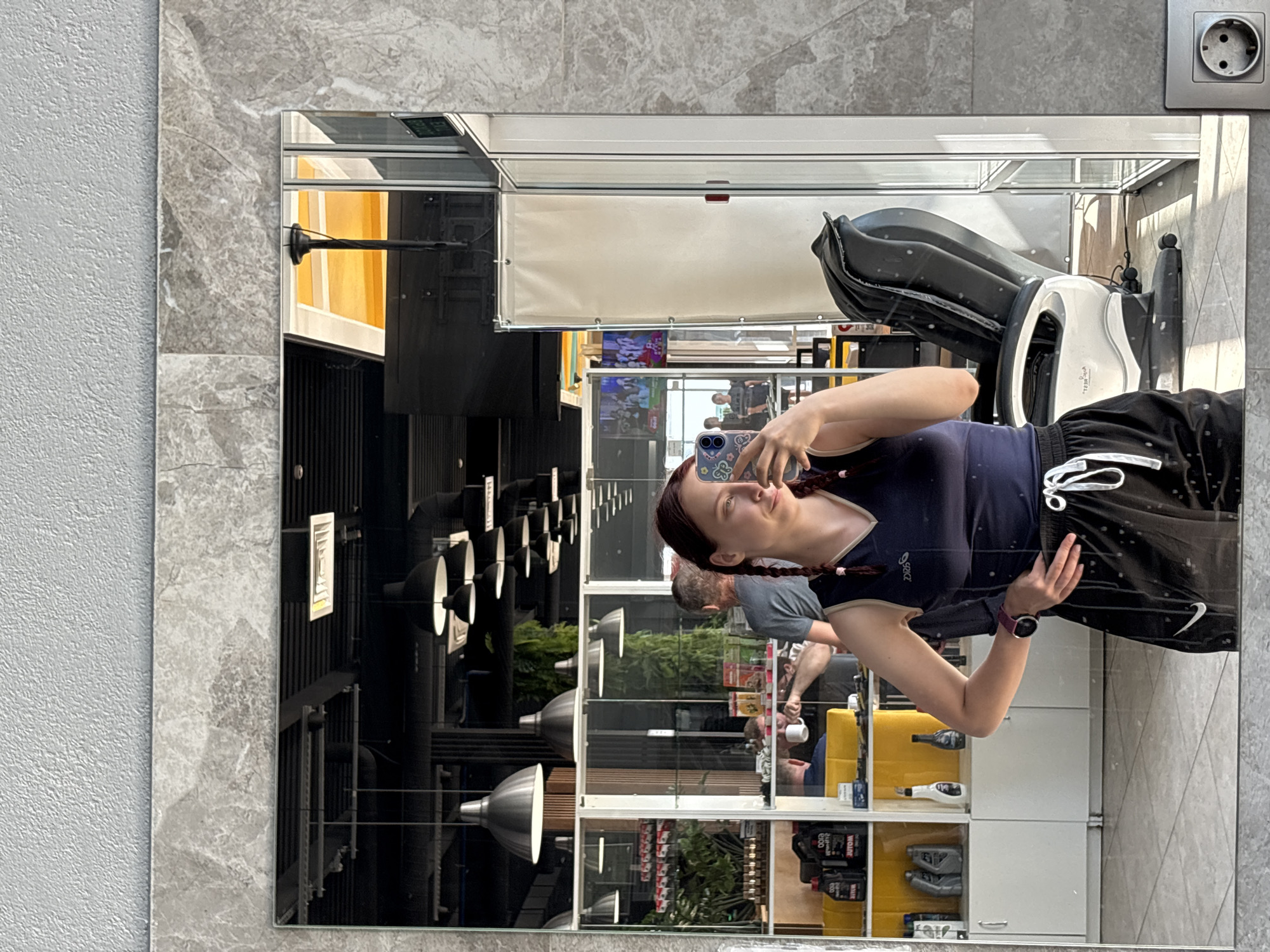 The image shows a reflection in a mirror. A woman is taking a selfie with her phone. She is wearing a blue shirt and black athletic shorts. A man in a gray shirt is partially visible behind her. The background reflects the interior of what appears to be a commercial establishment, possibly a shop. There are industrial-style lighting fixtures hanging from the ceiling, shelves with various products, and windows showing people outside. Based on the location of Казань, Russia, it might be a retail store, a gym, or a similar commercial venue.