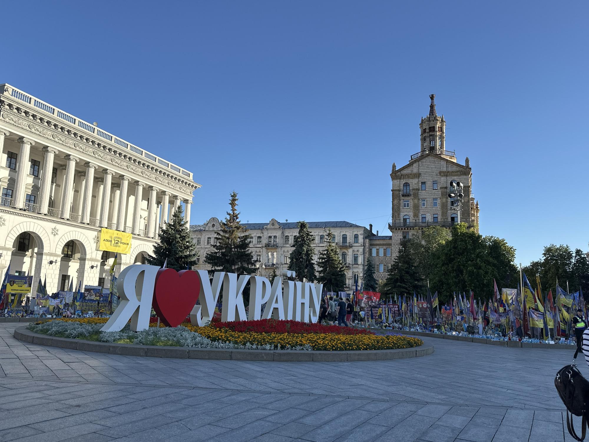 The image shows a wide-angle view of a public square in Kyiv, Ukraine, under a clear blue sky. In the foreground, a large, three-dimensional sculpture spells out "Я люблю Україну" (I love Ukraine) with a red heart replacing the letter 'o'. The sculpture is set within a circular flowerbed filled with red and yellow flowers. Behind the sculpture, a row of trees separates it from a line of Ukrainian flags and banners, many of which appear to be memorials or commemorative displays, arranged on the ground. Several people are visible in the background, some walking and others gathered near the flags.
To the left of the sculpture is a grand, white building with classical architecture, featuring tall columns and arched windows. Yellow banners are hung on this building. To the right and slightly further back, a tall, ornate building with a distinctive tower rises against the sky. This building features a clock face and numerous windows. In the mid-ground, other buildings with traditional architecture are visible, as well as more trees. The ground of the square is paved with large, grey stone slabs. A person is partially visible on the far right edge of the frame, with a bag. The overall scene appears to be a well-known public space in Kyiv, likely Maidan Nezalezhnosti (Independence Square), given the prominent "I love Ukraine" monument and the context of national pride and remembrance suggested by the flags and banners.