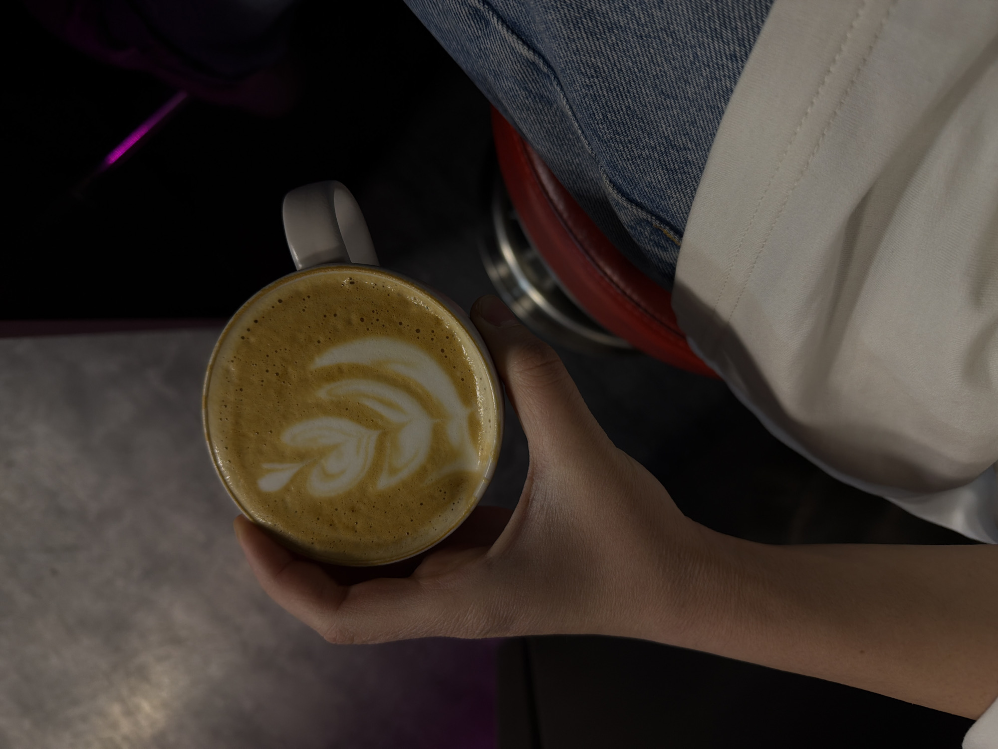 An overhead shot depicts a person holding a cup of coffee with latte art. The individual is seated, wearing blue jeans and a white top. A red stool is partially visible behind the person's leg. The coffee cup is held in the person's right hand. The background includes a metallic surface and a dark area with a hint of purple lighting. The setting appears to be a cafe or similar establishment. There is no evidence indicating it is in Yerevan, Armenia.