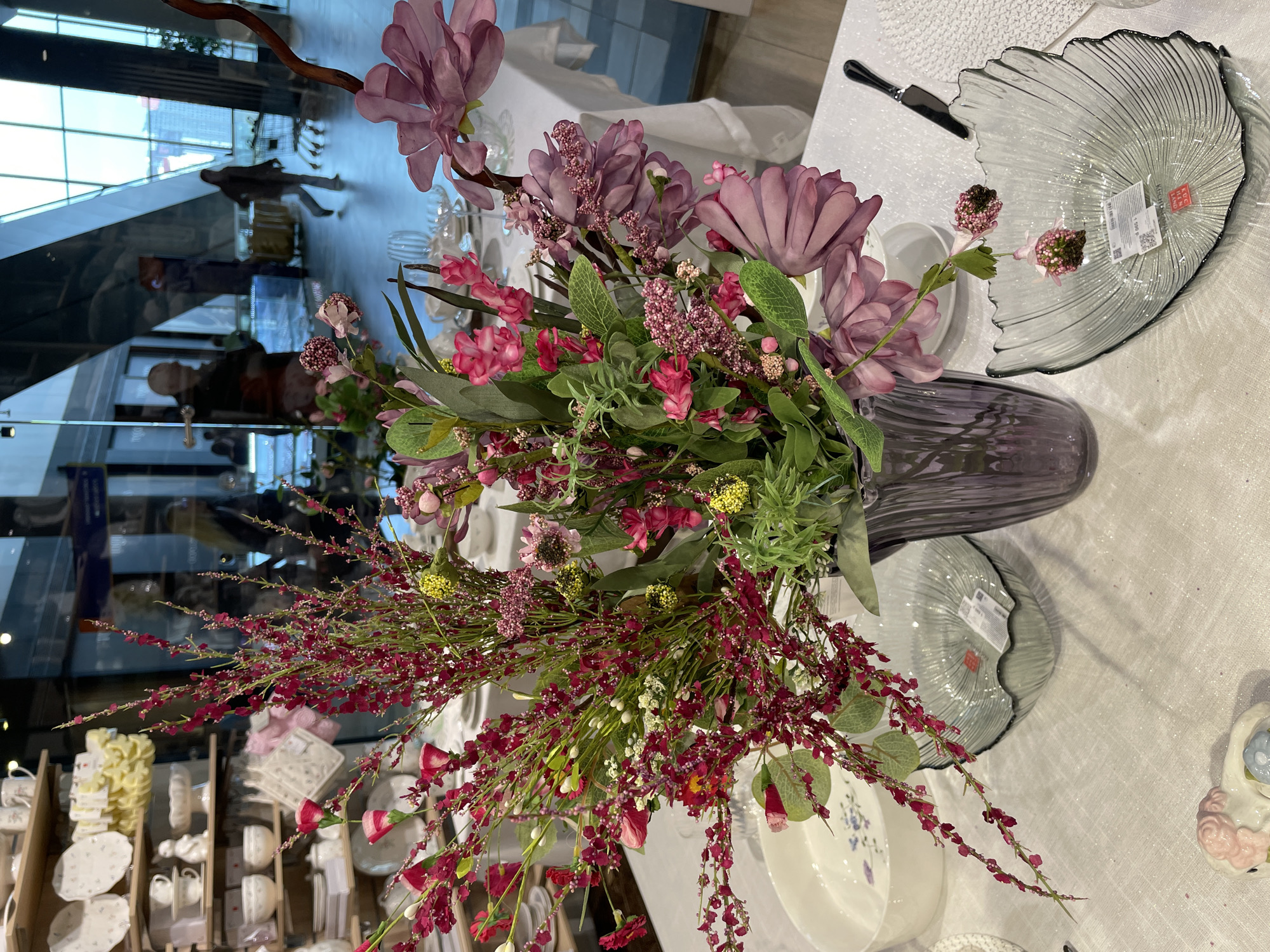 The image captures a floral arrangement in a retail setting in Kommunarka, Russia. The primary focus is a vase filled with pink, purple, and red flowers, displayed on a white tablecloth. The arrangement includes a variety of flower types and foliage, creating a dense, colorful display.
Several decorative objects are positioned around the floral arrangement, including ornate glass plates and various dishware sets. The background shows shelves stocked with additional tableware.
The reflections in the glass reveal parts of the surrounding environment.