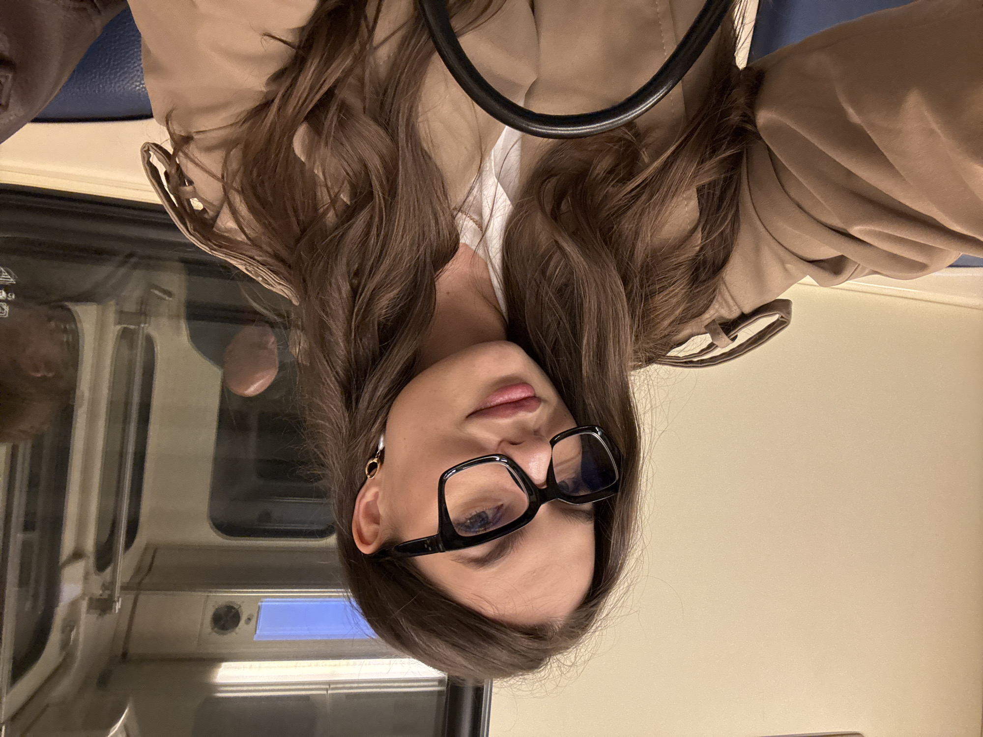 A young woman with light brown hair and wearing glasses is on a train, likely in Moscow, Russia. She is wearing a light brown coat or jacket. The background shows the interior of a train car, with windows, seats, and other passengers visible. The scene appears to be a casual snapshot taken during her commute or travel.
