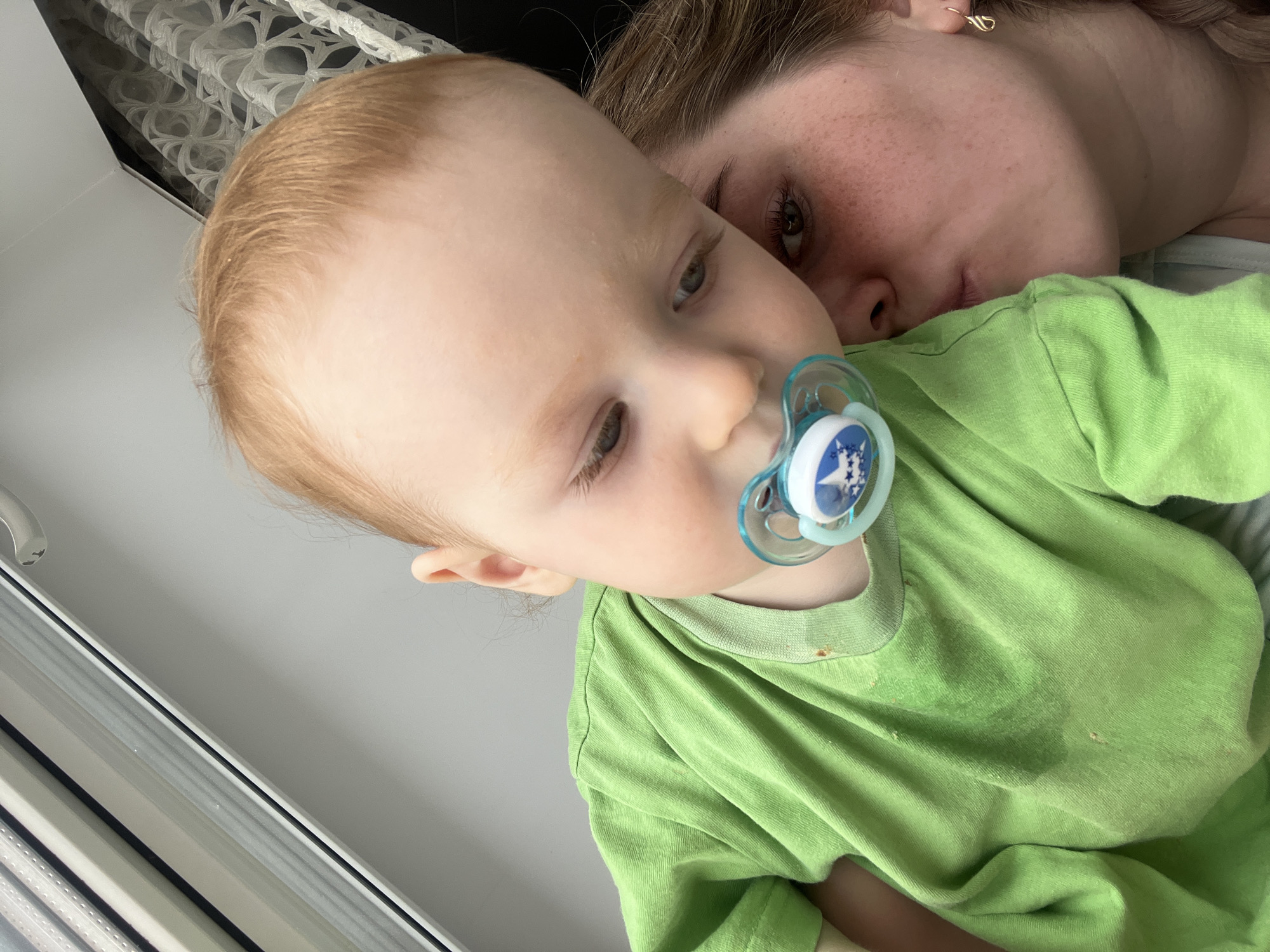 The image depicts an indoor residential scene in Chaikovsky, Russia, featuring an infant held by an adult female.
The infant, positioned centrally and angled slightly downwards, has fair skin, reddish-blonde hair, and light-colored eyes, gazing downwards and to the right. The infant wears a bright green, long-sleeved t-shirt with visible dampness and minor light-brown stains on the chest and around the neckline. A blue and white pacifier with a white star design is present in the infant's mouth.
The adult female is partially visible on the right side of the frame, comprising her right eye, cheek, and ear. She has fair skin with freckles across her cheek and brown hair, some of which appears braided or loosely gathered. A gold-colored hook earring is visible in her right ear. Her lips are pursed in a gesture directed towards the infant's side, suggesting a kiss. A light blue or striped garment belonging to the adult is partially visible beneath the infant's left arm.
The background consists of a white window frame with internal horizontal blinds on the upper left. The upper right shows a white wall meeting a dark surface, behind which a white patterned curtain with a floral or geometric design is visible. The scene is brightly lit by ambient light from an unseen source, consistent with an indoor setting.