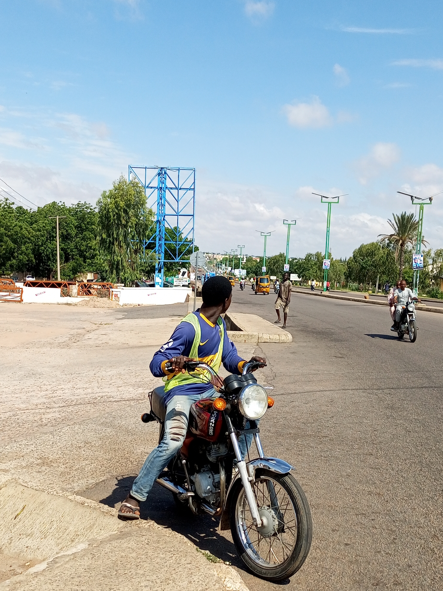 The image depicts a street scene in Tempure, Nigeria, under a bright, partly cloudy sky. In the foreground, a man wearing a blue long-sleeved shirt, a yellow and green reflective vest, and ripped blue jeans is seated on a red motorcycle. He is looking to his left, away from the camera. The motorcycle has a chrome front fender and headlight. The man's left foot is in a brown sandal.
The street is paved and appears to be a main road, with several other vehicles visible. To the right of the man, another motorcycle carries two people, a man driving and a woman as a passenger. Further down the road, a yellow three-wheeled taxi is seen. On the left side of the road, there is a sidewalk with a concrete curb. Beyond the sidewalk, there are trees and a large blue metal structure, possibly a billboard frame or part of a construction. Streetlights, several of which are solar-powered, line the right side of the road. In the background, there are more trees and buildings, suggesting an urban environment. The overall impression is a typical day in a Nigerian city with active street life and transportation.