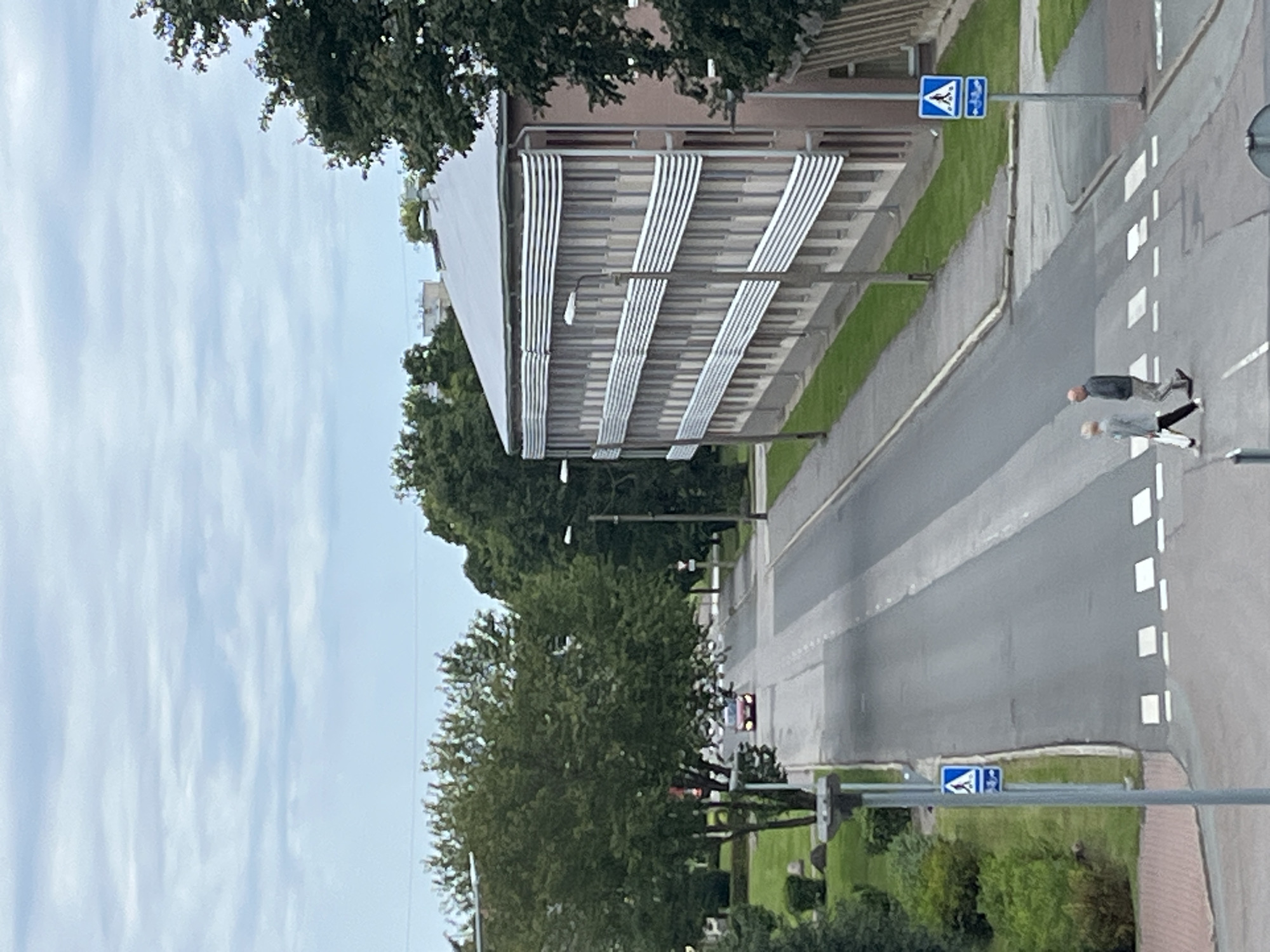 This is an aerial, medium-distance photograph taken in Kohtla-Järve linn, Estonia, on an overcast day. The scene depicts a street intersection with a crosswalk. Two individuals, an elderly man and woman, are walking across the street at the crosswalk. The man is on the right, wearing a dark jacket and light pants, while the woman is on the left, wearing a light jacket and dark pants. They are walking in the same direction, away from the viewer.
To the right of the street is a multi-story building with a light-colored facade and horizontal white accents. A pedestrian crossing sign is visible on a pole in front of this building. Trees line the left side of the street, and a red car is visible further up the road, traveling away from the viewer. The sky is filled with soft, horizontal cloud formations. The overall impression is a quiet, ordinary day in a town setting.