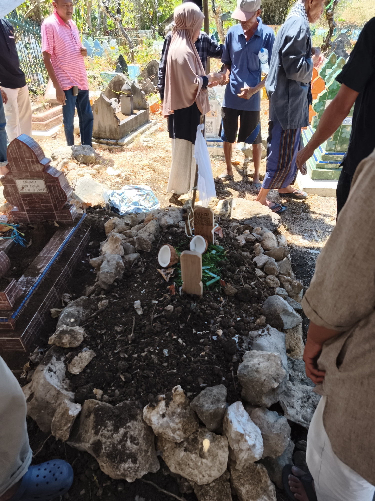 In the city of Tonasa, Indonesia, a group of people gathered at a cemetery. In the foreground, a freshly dug grave was surrounded by rocks and dirt. A half-coconut and a wooden marker with writing were placed on the grave. In the background, several other graves were visible, some adorned with colorful tiles. People dressed in traditional clothing were standing near the graves, some with their hands clasped. The cemetery appeared to be located in a dry, sunny environment with trees and vegetation in the distance.