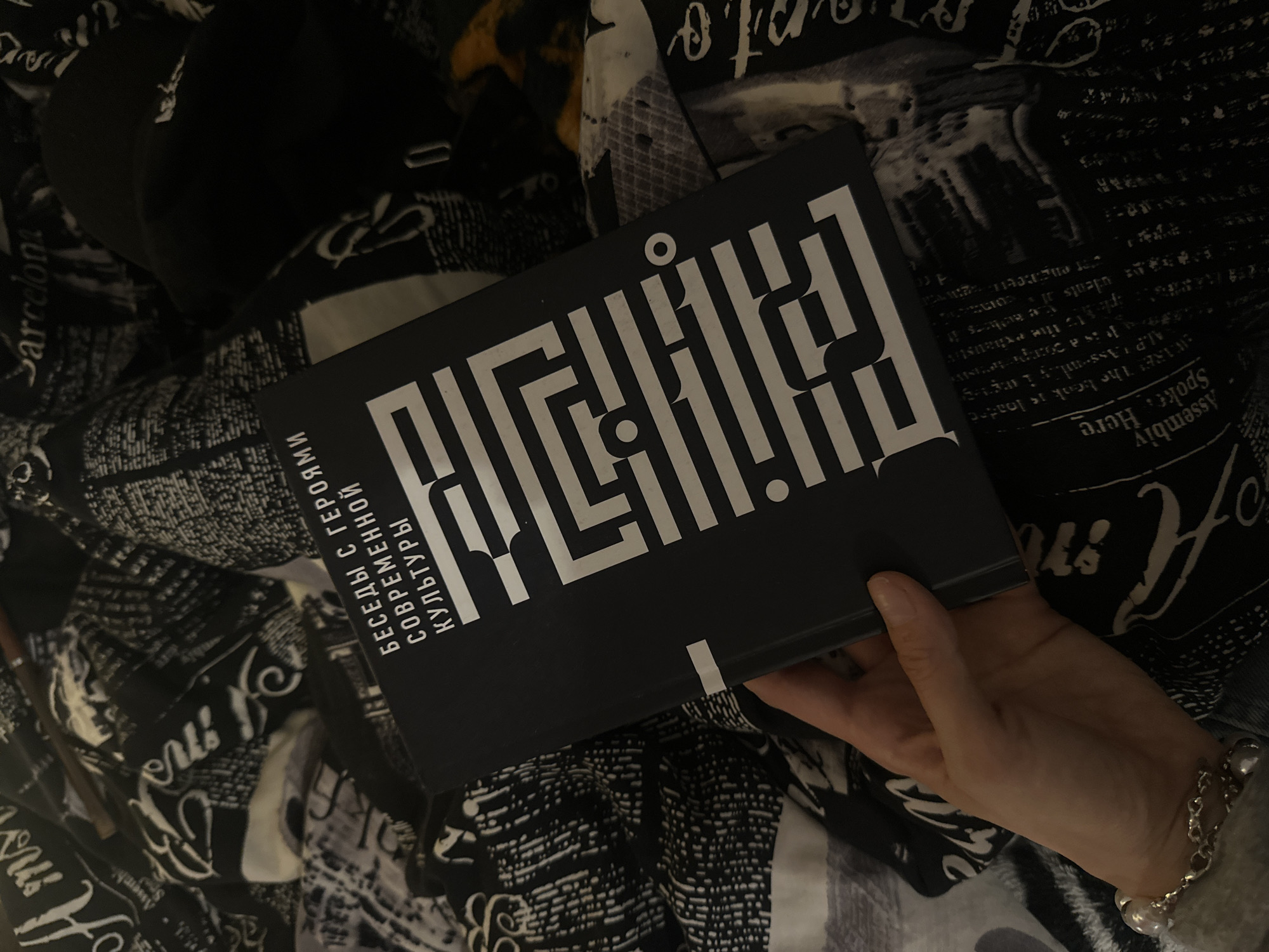 The image displays a close-up, top-down perspective of a single human hand holding a book, resting on a patterned fabric surface. The lighting is dim, indicating an indoor setting.
The subject's right hand, with light skin tone and well-maintained nails, is positioned in the lower left quadrant, gripping the bottom edge of a dark-covered book. A multi-strand bracelet adorns the wrist, featuring silver-colored chains interwoven with large, round, lustrous white beads and smaller metallic spheres.
The book has a matte black cover with white text and a geometric white graphic design. The prominent graphic on the right side of the cover forms the word "ЛАБИРИНТ" (Labyrinth) in a stylized, maze-like font. Above this, vertically stacked in smaller white sans-serif text, are the phrases: "БЕСЕДЫ С ГЕРОЯМИ" (Conversations with Heroes), "СОВРЕМЕННОЙ" (of Contemporary), and "КУЛЬТУРЫ" (Culture). A narrow horizontal white stripe is visible on the book's spine area, near the bottom.
The book and hand are resting on a patterned bedspread or blanket, primarily in black and white tones. The pattern consists of various architectural sketches, cityscape outlines, and textual elements. Visible English words include "Amsterdam" at the top left, "Barcelona" at the top right, "Assembly Here" (partially) at the bottom right, and "arts &" (partially) also at the bottom right. Other script-like and abstract patterns are also present across the fabric.
In the upper central background, a thin, dark, cylindrical object, possibly a pen or small stick, lies horizontally on the patterned fabric. To the upper right, a black fabric object, resembling a cap or hat, is partially visible. Further to the far right, a small portion of a textured, yellowish-orange and black patterned fabric or object is discernible.