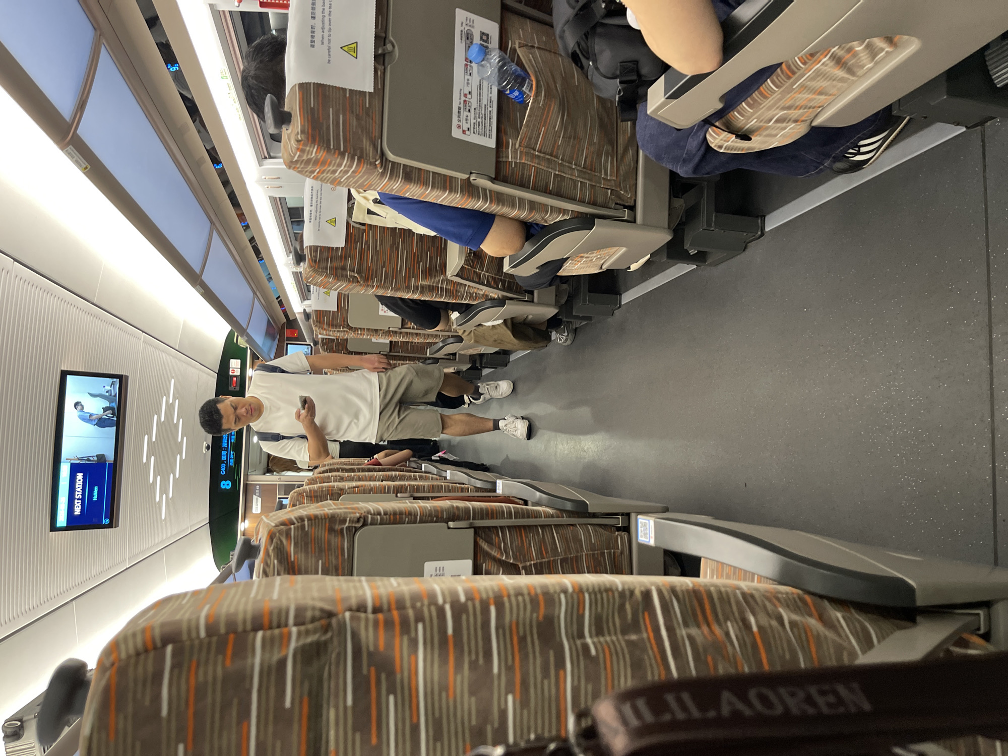 This image captures the interior of a train, likely a high-speed rail service in China, given the "Longhua District, China" context. The perspective is from a passenger's seat, looking down the aisle. The primary subjects are the seats themselves, upholstered in a brown fabric with vertical stripes of white and orange. A man in a white t-shirt and grey shorts stands in the aisle, holding a smartphone, and looking down at it. He is wearing white sneakers. Other passengers are visible in the background, seated in the train cars. One passenger is seen with their legs extended, and another has their arm resting on the back of a seat.
A prominent object in the scene is a screen mounted on the wall of the train, displaying "NEXT STATION HuMen". There are also safety instructions printed on the back of the seats, one of which reads, "调整椅背时,谨防傾倒 when adjusting the back be careful not to tip over the tea". This suggests the train is in motion, and passengers are being reminded of safety procedures. The lighting appears to be artificial, consistent with an indoor environment like a train car. The overall scene suggests a typical journey on a public transportation system, with people engaged in their own activities while traveling. The presence of the man with his phone and the overhead lights imply it could be daytime, though no external weather cues are visible. The text on the screen and the safety instructions provide specific details about the location and operational aspects of the train.
