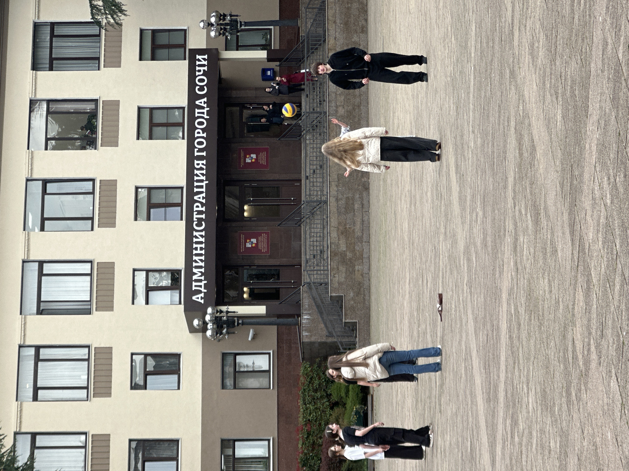 The image depicts a public square in front of the City Administration building in Sochi, Russia. The building, identified by the sign "АДМИНИСТРАЦИЯ ГОРОДА СОЧИ," is a multi-story structure with a light-colored facade and dark-framed windows. Its main entrance, featuring dark double doors, is accessed via a set of stone stairs with dark metal railings. Two multi-lamp streetlights with reflective spherical tops flank the entrance, and a blue mailbox is visible to the right of the stairs.
In the foreground, on the paved square, seven young individuals are present. Five of them are actively engaged with a yellow and blue volleyball, which is airborne between them. A female with long light hair, wearing a white puffer jacket and dark pants, has her arms slightly outstretched. To her right, a male in a black tracksuit observes the ball. Another female, partially obscured, is behind the first, with raised arms. Two more females stand to the far left, one in a white top and dark pants, the other in a dark top and dark pants, facing the group. Two additional individuals are visible on the building's entrance stairs, observing the scene. A brown bottle lies horizontally on the paved ground in the mid-left area. The ground consists of greyish-brown paving stones. The sky appears overcast.
