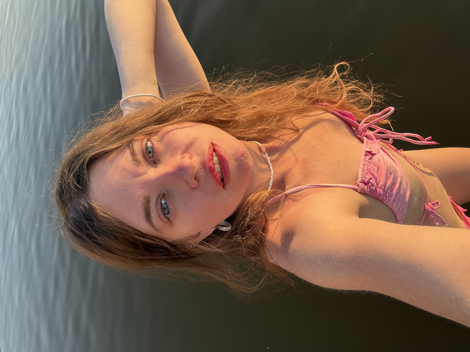 A woman with long, auburn hair is lying on her back in dark water, possibly a lake or river, with ripples visible on the surface. She is wearing a pink polka-dot bikini. Her arms are raised above her head, and her left hand is resting on her forehead. She has blue eyes and is looking up at the camera with a gentle expression. A faint heart tattoo is visible on her left arm. She is wearing a delicate pearl necklace and a shell earring. The lighting suggests it is late afternoon or early evening, with the sun casting a warm glow on her skin. The scene appears to be a serene and personal moment, perhaps during a summer swim in Sol-Iletsk, Russia.
