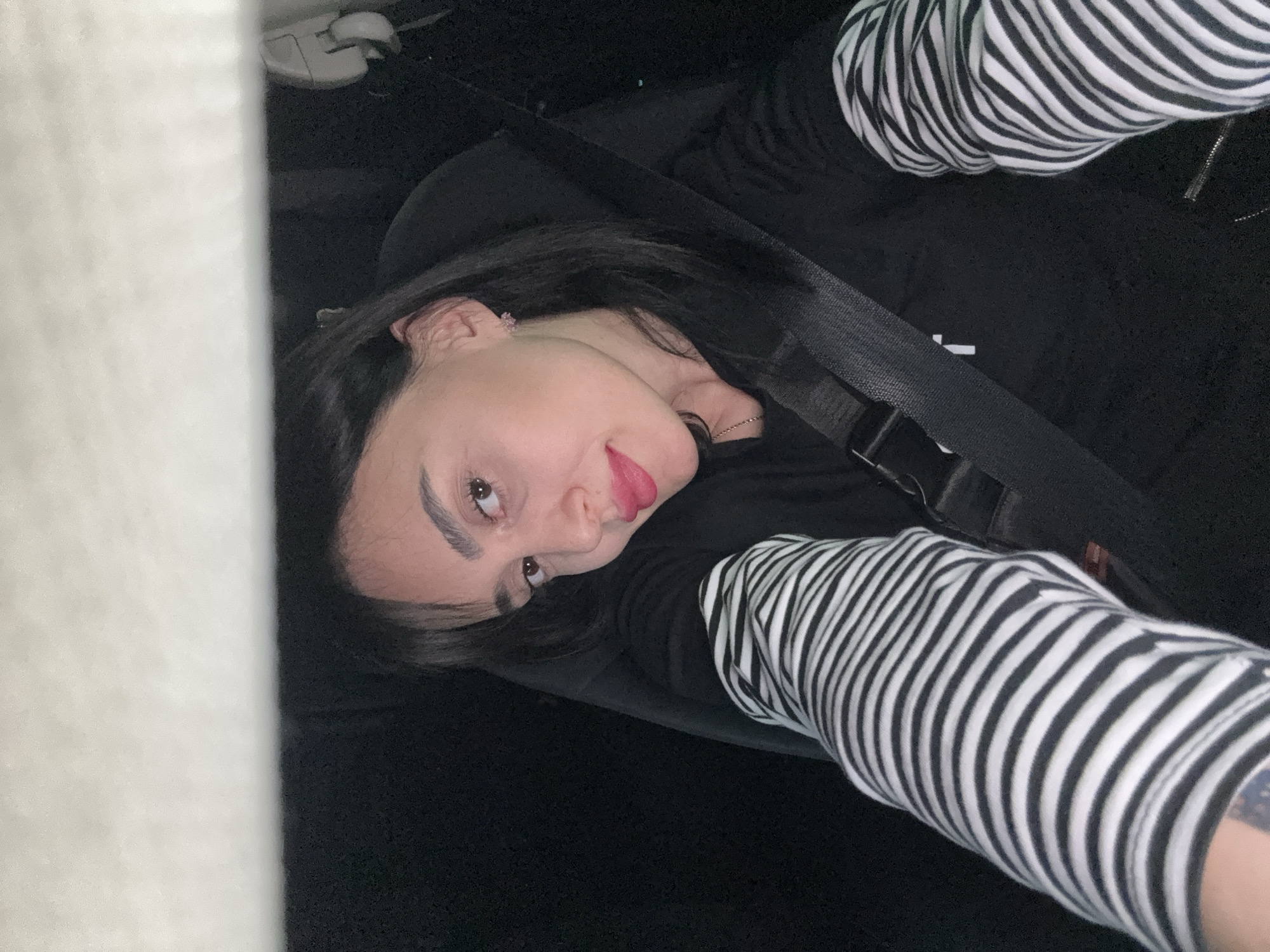 A young woman is captured in a close-up portrait, oriented sideways, inside what appears to be a vehicle. Her head is tilted, and she is looking upwards and to the left with a slight, subtle smile and red lipstick. She has long, dark hair, neatly defined eyebrows, and a small earring is visible in her right ear. A thin silver chain necklace is also visible.
She is dressed in a black top with a black and white striped long-sleeved garment underneath, visible on both arms. A black car seatbelt crosses diagonally over her chest, secured in place. The interior of the vehicle is dimly lit, suggesting an evening or night setting. In the upper left background, part of the car's interior, possibly a headrest or a control panel, is dimly visible. A small, partially obscured white letter, potentially a 'T', is visible on her black shirt beneath the seatbelt. The overall scene suggests a casual moment, possibly a selfie taken while traveling in Tbilisi, Georgia.