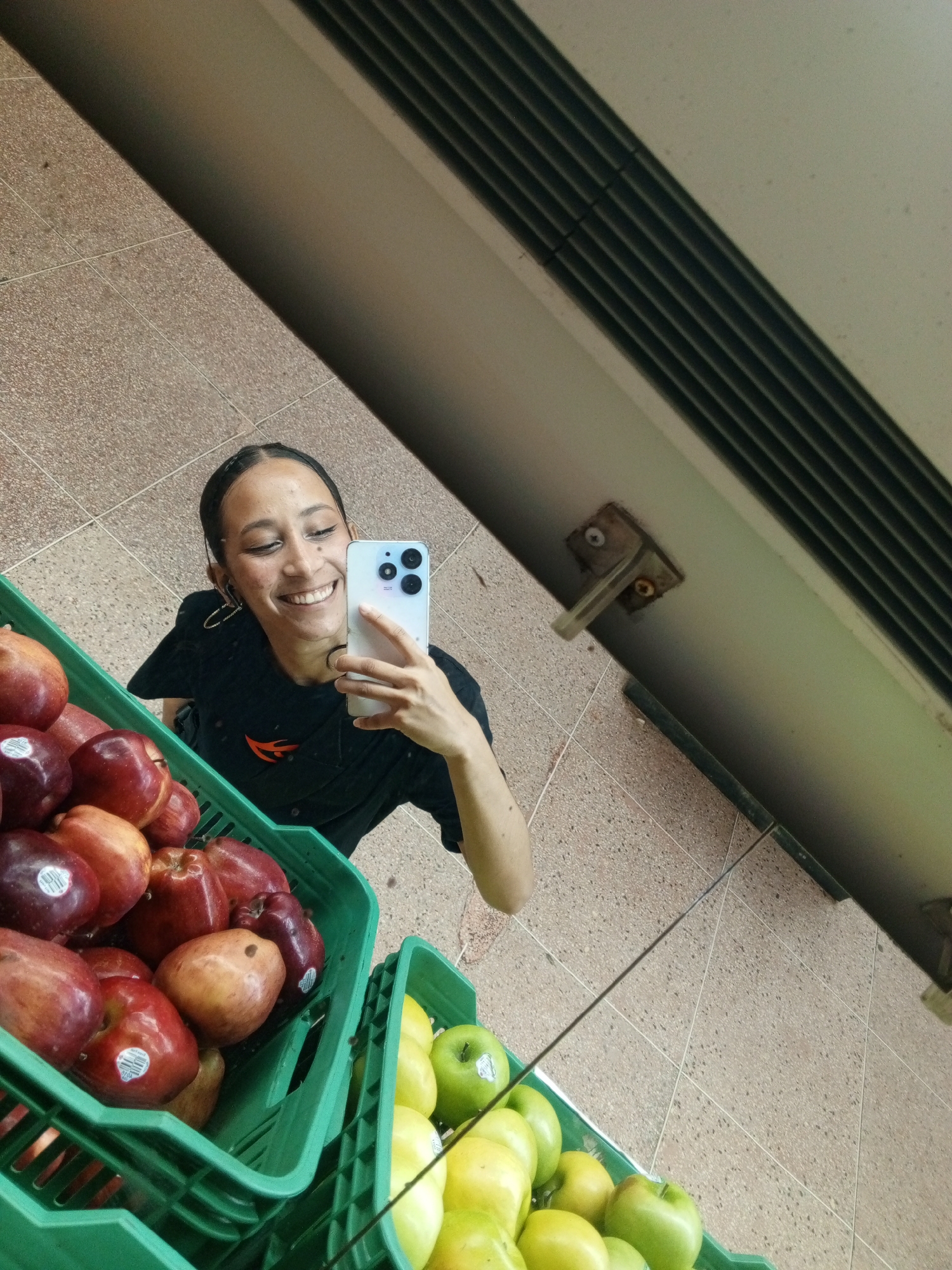 A young woman is holding a smartphone and smiling into the camera, which is positioned to take her picture in a reflective surface (likely a mirror) inside what appears to be a store. She stands next to two stacked green crates filled with apples; the top crate contains red apples, and the bottom crate contains a mix of yellow and green apples. The surface upon which the woman stands is tiled. The image seems to have been taken at an angle facing upwards to the right. Above her reflection are elements of the store's infrastructure: a grey panel with a ventilation grate. It is not possible to determine that the picture was taken in Porlamar, Venezuela from the given information in the image.