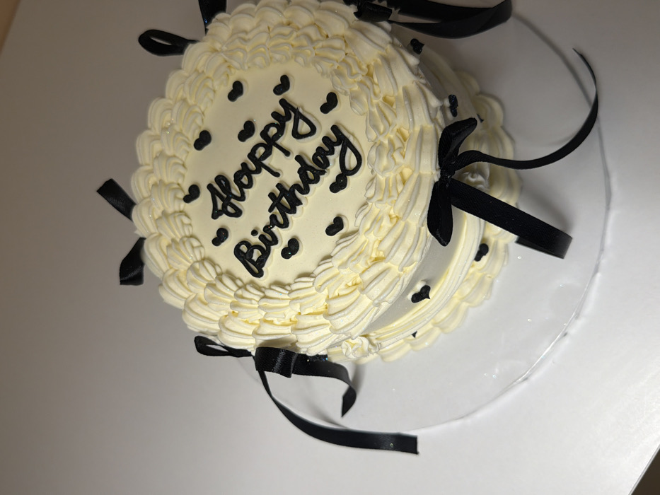 A two-tiered round cake, decorated with creamy white frosting and adorned with black ribbon bows, sits on a clear plastic cake stand. The top tier features "Happy Birthday" in black script, punctuated by small black heart decorations. Similar hearts are sparsely scattered on the lower tier. The frosting is swirled and ruffled, creating a delicate texture around the edges of both tiers. The cake's presentation is simple yet elegant, contrasting the light icing with the dark ribbons and lettering.