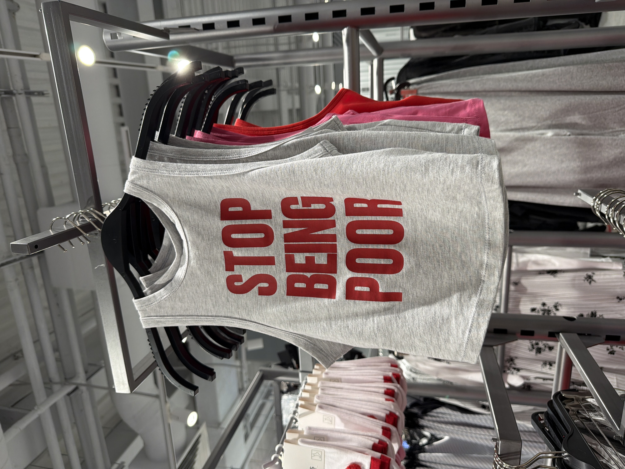 The image shows a rack of grey tank tops with "STOP BEING POOR" printed in red. The tank tops are hanging on black hangers from a metal rack in what appears to be a retail store. Other clothing items, including socks and more tank tops of different colors, are visible in the background. The overall environment is clean and well-lit, suggesting a commercial setting. There is no information in the image to suggest a specific location in Moscow, Russia.