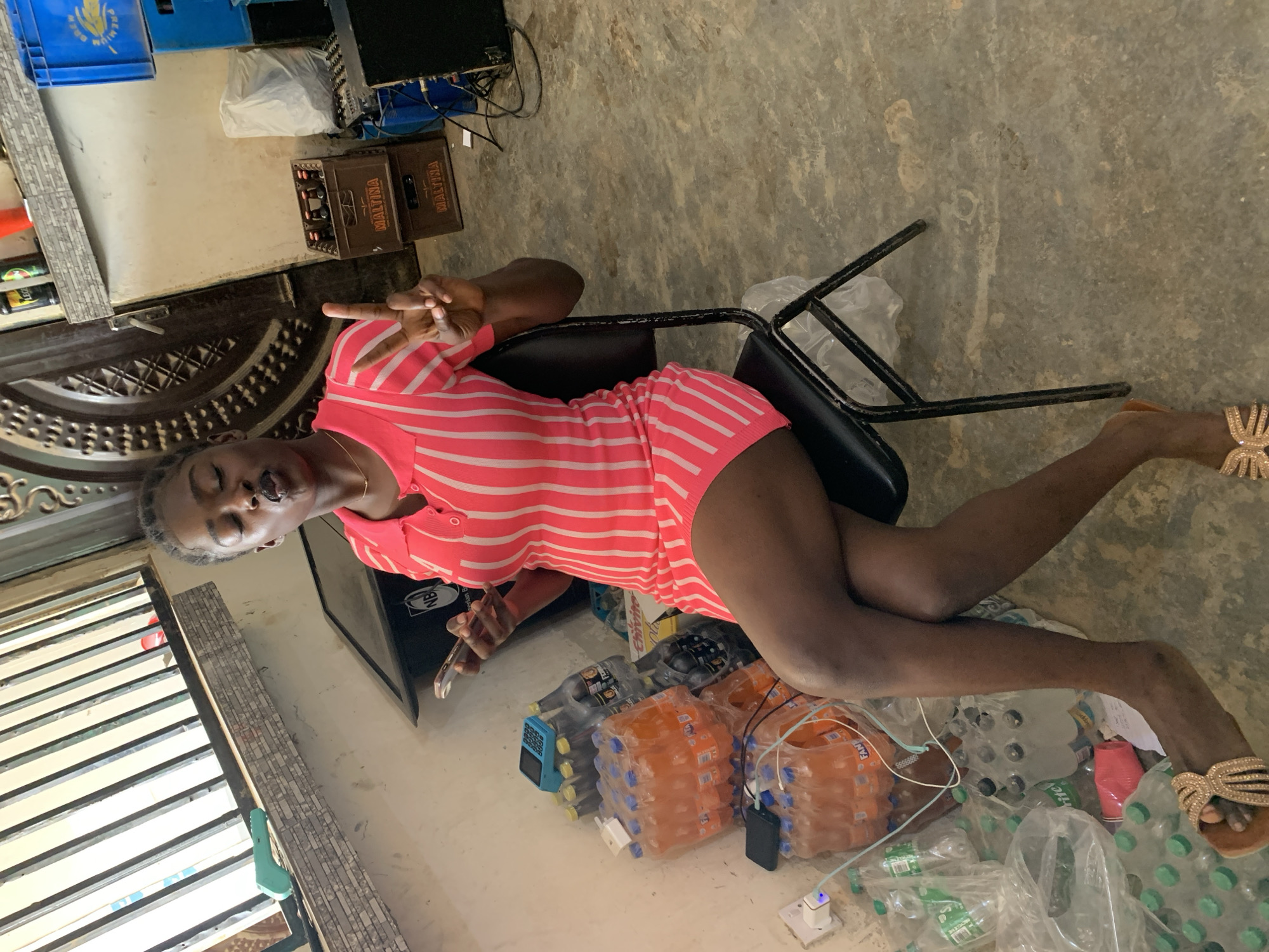 The image depicts a woman sitting on a black chair indoors. She is wearing a short, horizontally striped pink and white dress and tan sandals. She is holding her phone and making a peace sign with her left hand. She has short, braided hair. The room appears cluttered, with stacks of plastic water bottles, crates, and other items on the floor. The location is identified as Jos, Nigeria.