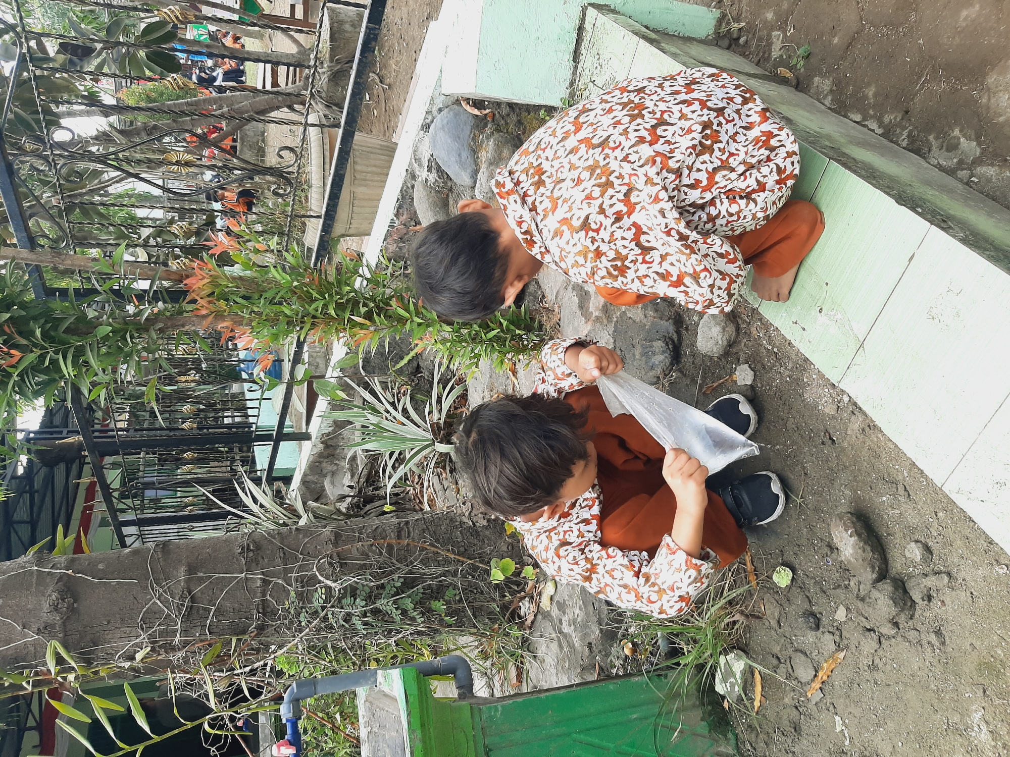 Two young boys, dressed in matching patterned shirts and orange pants, are crouched down in a schoolyard in Sumberjo, Indonesia. The boy on the left is examining a clear plastic bag filled with water. The boy on the right is also looking at the bag, his right hand extended towards it. They are surrounded by dry dirt, scattered rocks, and sparse vegetation. In the background, a black metal fence encloses a courtyard with trees and other children sitting on the ground. The scene appears to be outdoors during daylight.