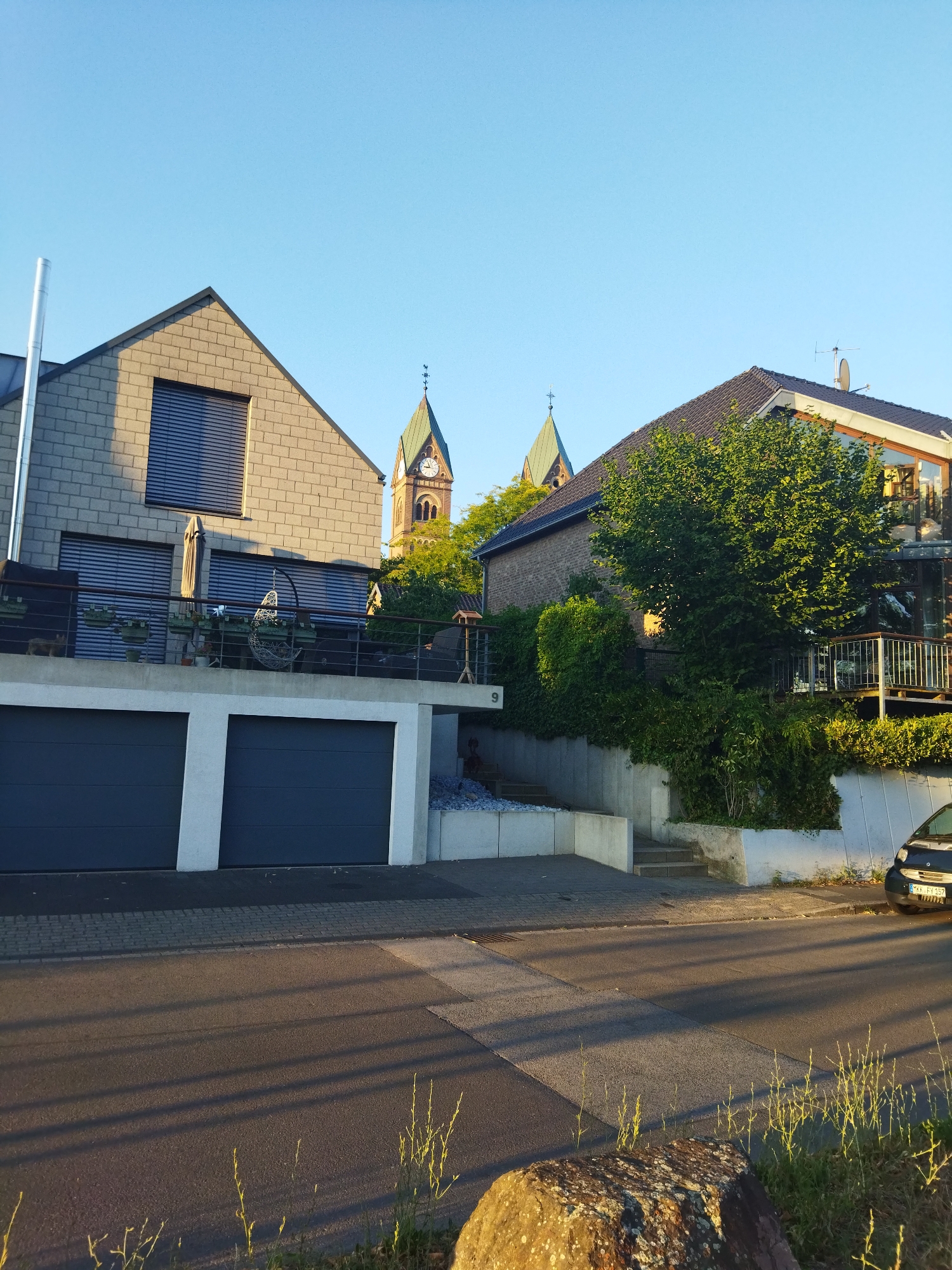 In Leverkusen, Germany, on a clear day with blue skies, a street scene unfolds. In the foreground, a paved road with distinct lane markings is illuminated by long shadows cast by unseen objects. A large, textured rock sits on the right side of the road, partially obscuring a patch of dry grass.
Behind the road, a modern house with a gray brick facade and a dark-tiled roof dominates the left side of the image. It features two large, dark gray garage doors on the ground level. Above the garage, a balcony or terrace is visible, furnished with outdoor seating and potted plants, accessible by a sleek metal railing. A metal chimney pipe extends vertically from the left side of the house.
To the right of this house, a second, older brick house is partially visible, with a dark-tiled roof and lush green foliage surrounding it. A large tree with dense foliage is situated in front of this house. A balcony with a metal railing is also present on this property, and large glass windows are visible on its upper level.
In the background, the spires of a church or cathedral rise above the houses. One spire is topped with a cross and features a clock face. The spires are a muted green color and appear to be constructed of stone. The overall impression is one of a suburban residential area with a blend of modern and older architecture, set against a backdrop of clear skies and greenery.