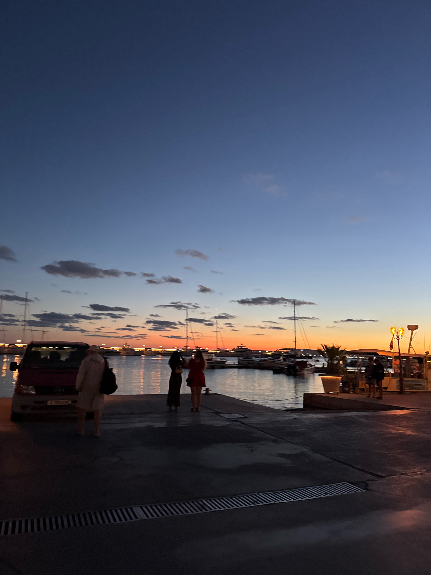 The image captures a twilight scene at a marina in Sveti Vlas, Bulgaria. The sky transitions from a deep blue at the top to a vibrant orange and yellow near the horizon, indicating sunset or sunrise. Scattered clouds are visible in the sky. Several boats and yachts are docked in the water, their reflections shimmering on the surface.
In the foreground, on the concrete pier, a red van is parked on the left. Three individuals are standing near the van. One person, wearing a light-colored jacket and shorts, is facing away from the camera. Two other figures, dressed in dark clothing and a red dress respectively, are standing together, appearing to interact with each other. Further down the pier, a couple of individuals are visible near a brightly lit structure. The ground in the foreground is a dark, wet-looking concrete with a metal grate running across it.