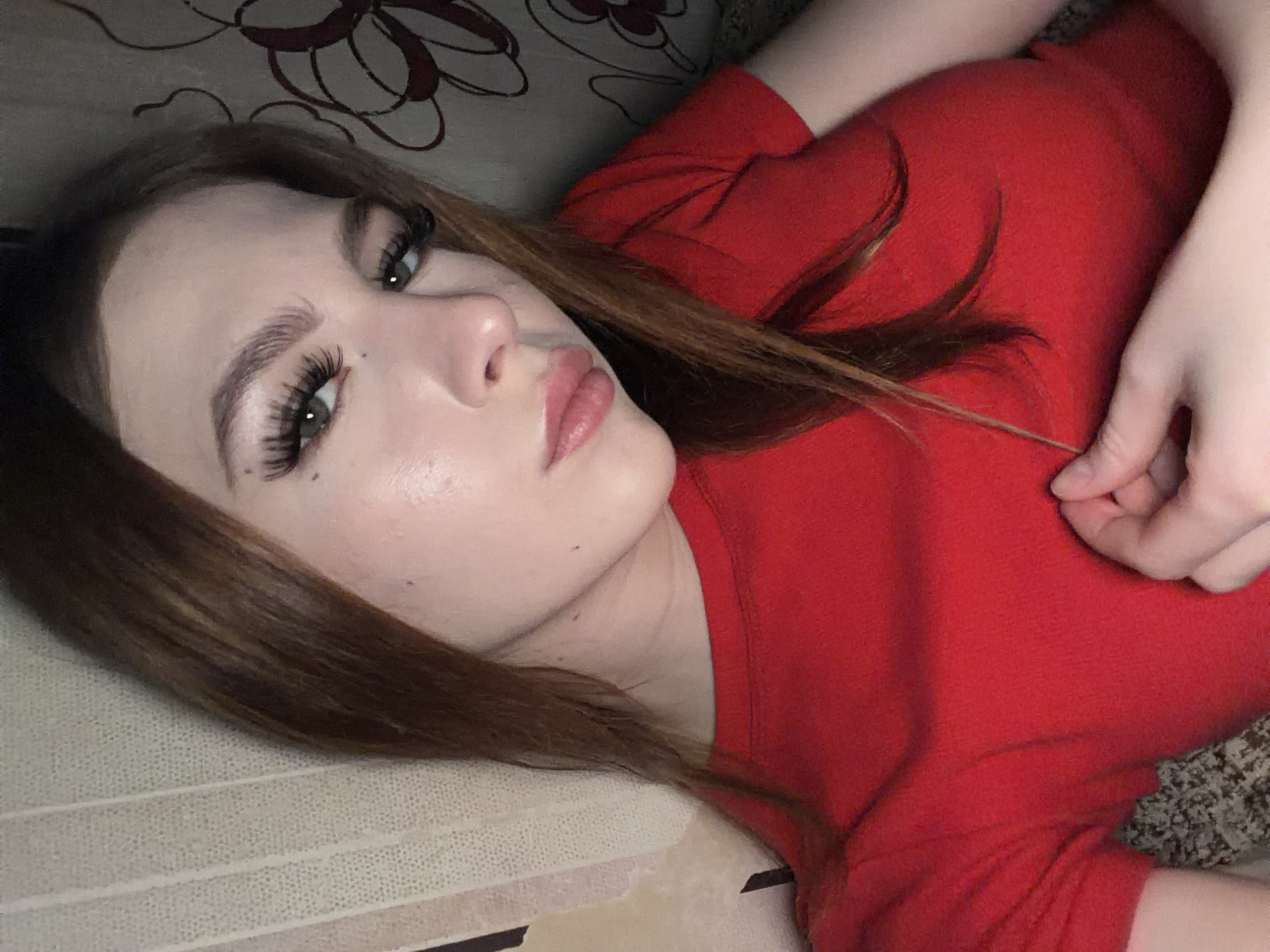 A fair-skinned woman with auburn hair and long eyelashes is lying down. She wears a red shirt. The background includes patterned surfaces, possibly a couch or bed. The specific location is not discernible from the image.