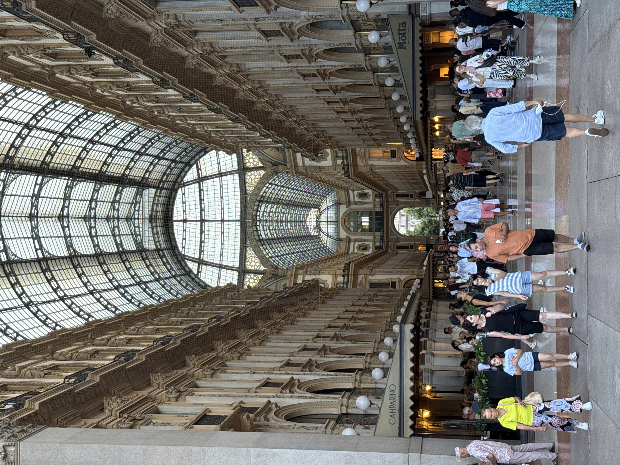The image depicts the Galleria Vittorio Emanuele II in Milan, Italy, a historic shopping arcade with a distinctive glass and iron roof. The perspective is a low-angle, vertical shot capturing the grandeur of the architecture and the activity within the arcade.
In the foreground, several individuals are walking or standing. A woman in a bright yellow shirt and patterned pants is in the lower left, holding a beige bag. Next to her, a young girl in a blue t-shirt and shorts stands with her hands clasped. Further to the right, a woman in a black top and shorts walks past, followed by another woman in a striped t-shirt and denim shorts, eating. An older man in an orange t-shirt and black shorts is walking to the right. In the middle ground, a man in a blue button-down shirt and dark shorts walks away from the camera, holding a white mask.
The sides of the arcade are lined with ornate, multi-story buildings featuring arched windows, balconies, and decorative stonework. Signs for "CAMPARINO" and "Motta MILANO 1928" are visible on awnings above storefronts, indicating the presence of businesses. The ceiling of the arcade is a vast, arched glass and metal structure that allows natural light to filter through, creating a dramatic effect. People are seen milling about, dining at outdoor tables beneath the awnings, and shopping. The floor is paved with a mosaic of colorful tiles. The overall scene conveys a sense of bustling urban life within a remarkably elegant and historic setting.