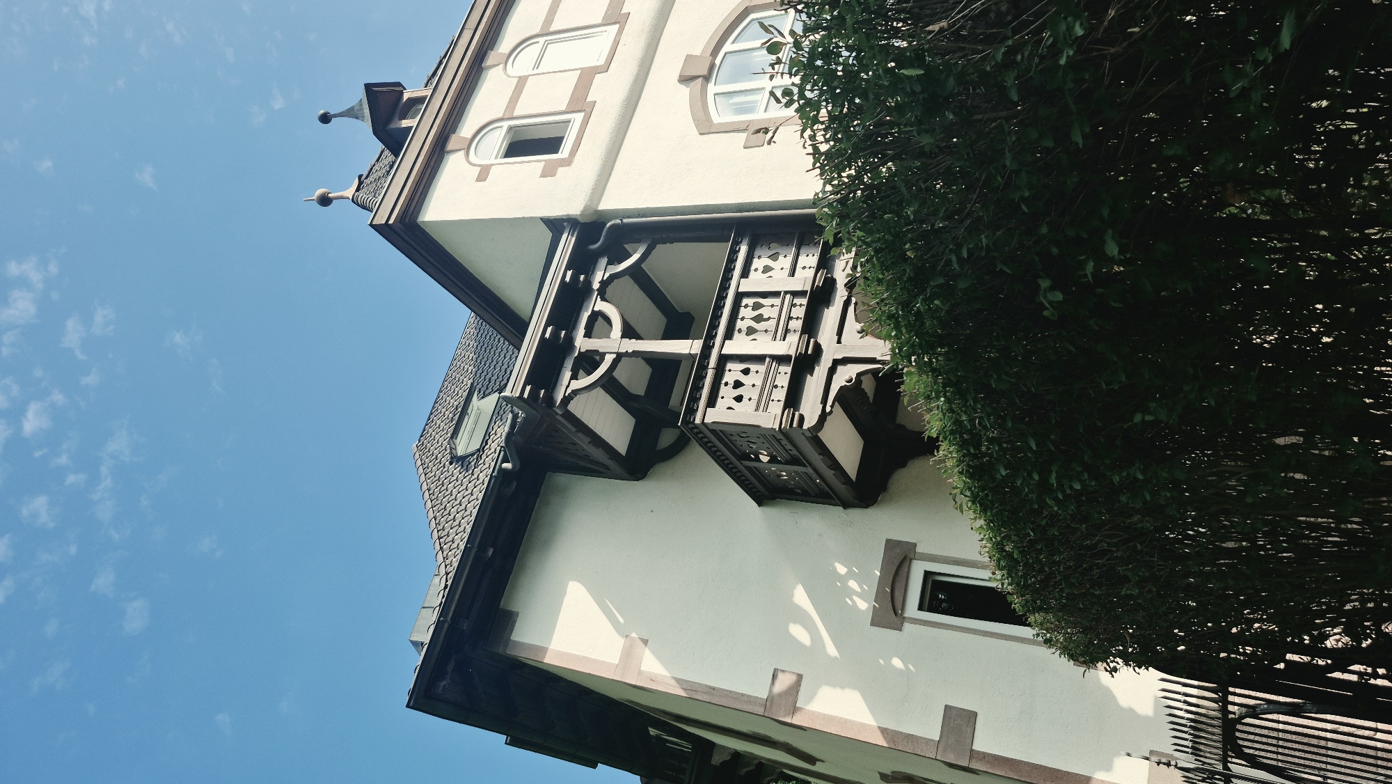 The image shows a low-angle view of the exterior of a building in Baden-Baden, Germany, against a bright blue sky with scattered white clouds. The building has a light-colored stucco facade with brown trim around windows and corners. A dark wooden balcony with ornate carvings and heart-shaped details extends from the upper floor. The roof is tiled with a dark color, and there are dormer windows visible. In the foreground, a dense, dark green hedge obscures the lower part of the building and a black metal fence. The lighting suggests it is daytime, possibly late morning or early afternoon, with shadows cast on the building.