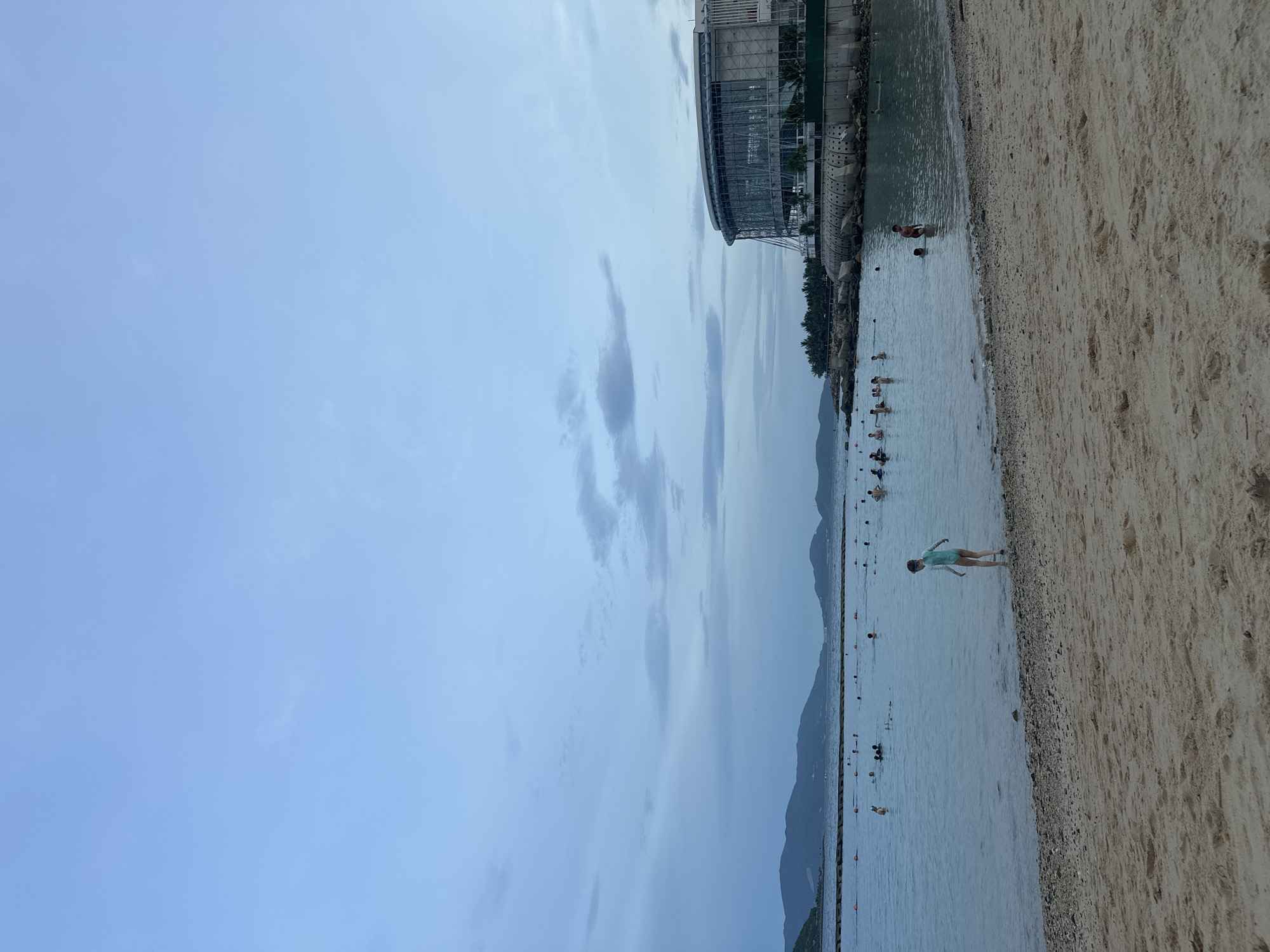 The image depicts a beach scene in Nha Trang, Vietnam, likely at Vinpearl Land. The sky is overcast with a pale blue hue, and scattered clouds are visible. In the foreground, a sandy beach stretches across the lower portion of the frame. A single child wearing a turquoise swimsuit stands near the water's edge, facing away from the camera.
The sea is calm and a light blue color. Numerous people are swimming and wading in the water, with some forming small groups. Orange buoys are present in the water, marking a swimming area. In the background, a large, modern, circular building with a glass facade is prominently situated on the right side of the image, overlooking the water. This building appears to be part of a resort or entertainment complex. Beyond the water and to the left, a line of hills or mountains can be seen in the distance, some appearing green and others shrouded in a hazy blue. The overall atmosphere suggests a late afternoon or early evening setting.