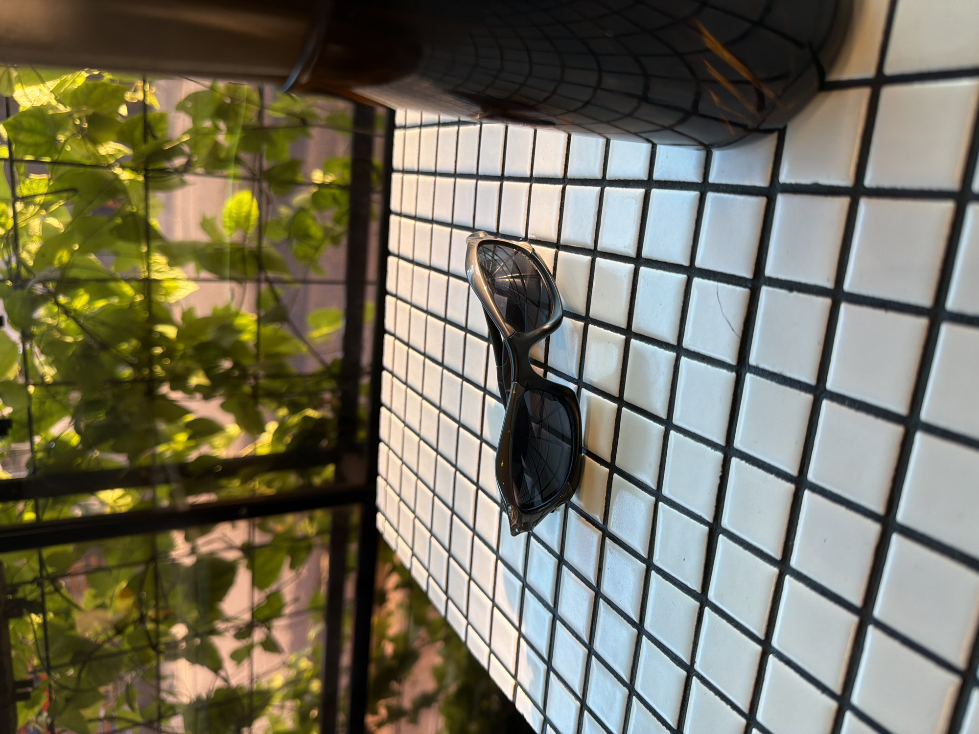 The image shows a pair of black sunglasses lying on a white tiled surface with black grout. A dark, reflective object (possibly a container) is partially visible in the upper portion of the frame. The background features a green leafy structure, suggesting an outdoor setting. Considering the context of North Kuta, Indonesia, the tiled surface may be part of a table at an outdoor cafe or restaurant with nearby greenery. There are no people visible in the image. The sunglasses are the central object of focus. No actions or interactions are evident.