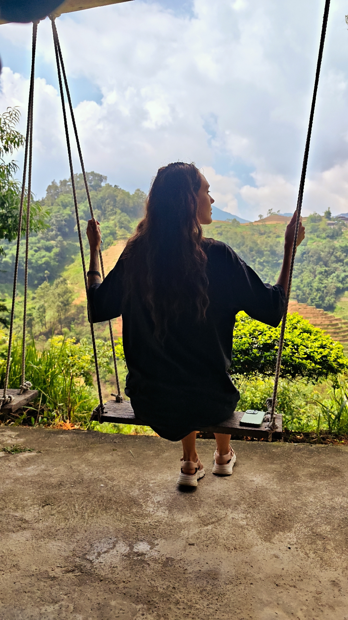 A woman with long, wavy dark hair is sitting on a wooden swing in Hoàng Su Phì Commune, Vietnam. She is facing away from the camera and looking to the right, towards the background. She is wearing a black, oversized t-shirt and light-colored sandals. The swing is made of wood and hangs from thick ropes. The ground beneath the swing is concrete. The background features a lush, green mountainous landscape with terraced rice fields and vegetation. The sky is partially cloudy. A smartphone is resting on the swing beside the woman.