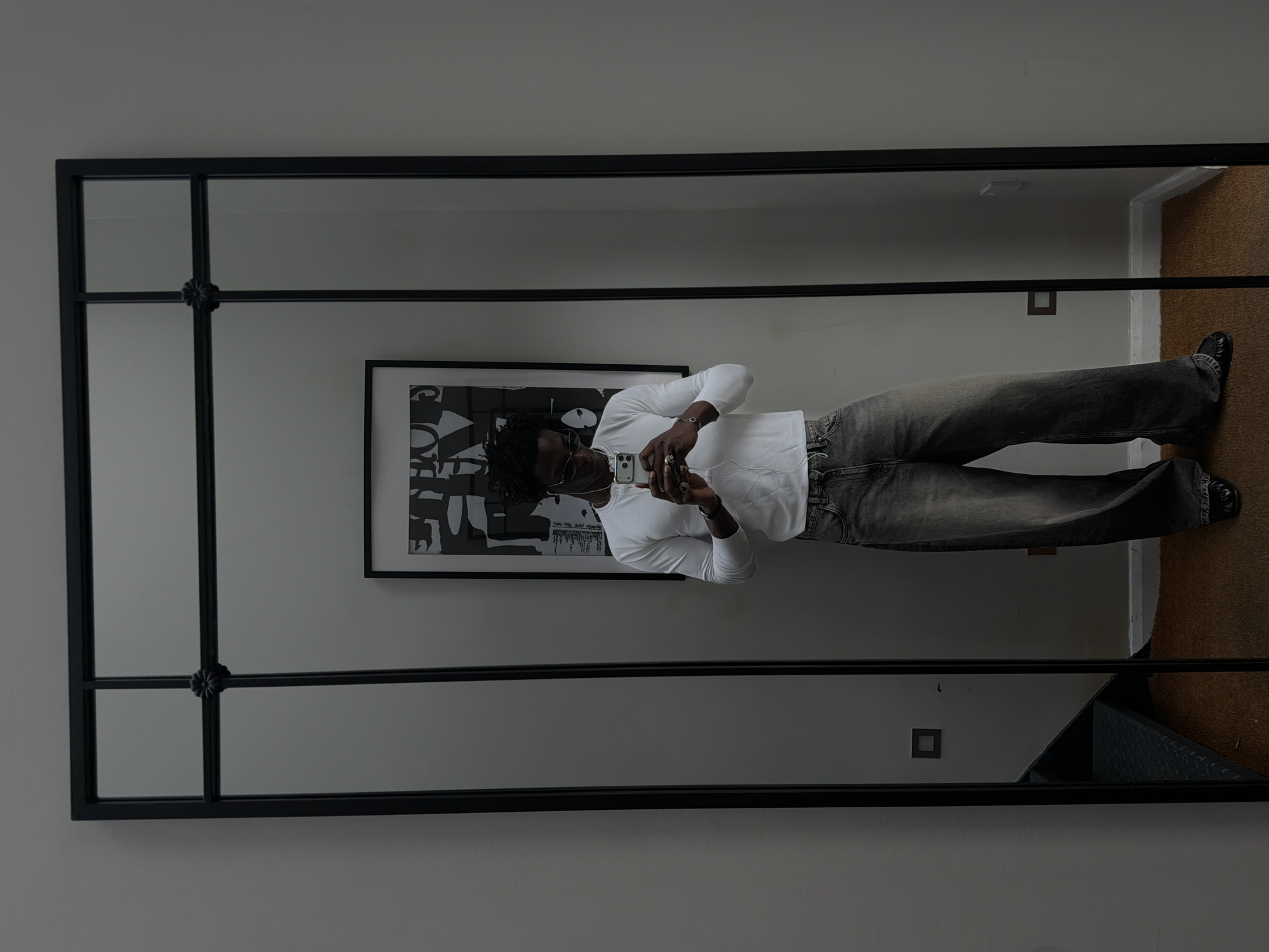 An individual is captured in a full-length mirror selfie, positioned indoors against a light grey wall. The mirror features a black metal frame divided into a grid pattern with a decorative floral-like rosette at the central horizontal and vertical intersection. The subject, a male with dark curly hair and sunglasses, is wearing a long-sleeved white t-shirt, baggy washed-out dark grey jeans, and dark shoes. He is holding a white smartphone with both hands, looking down at the screen, with white wired earphones extending from the phone. Multiple rings adorn his fingers, and a dark-colored bracelet or watch is on his left wrist. Reflected behind the individual is a framed piece of abstract black and white artwork. The room has light grey walls and a brown carpeted floor. Visible in the reflection on the left is a dark staircase with metallic treads and a square light switch, while on the right, another light switch is seen near the baseboard. The scene is set in London, United Kingdom.
