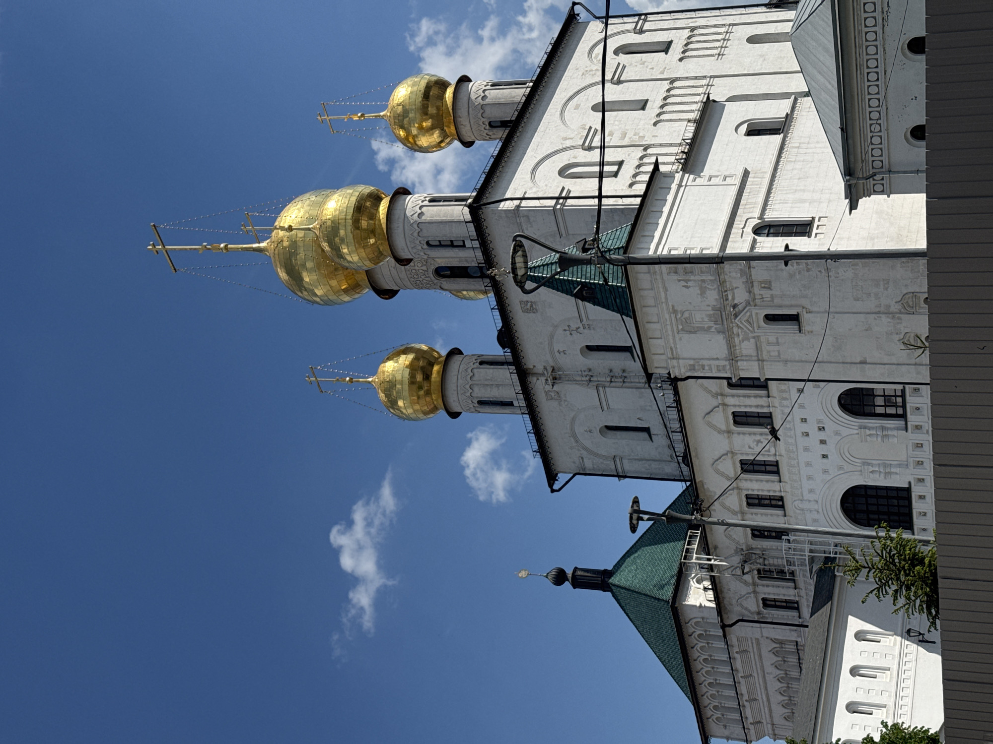 The image displays a majestic architectural complex featuring several white-walled buildings against a clear blue sky. Dominating the upper portion of the scene are three prominent golden, onion-shaped domes, each topped with an ornate Orthodox cross. Delicate chains extend from the crosses down to various points on the domes.
The main buildings are constructed of white stone, exhibiting intricate architectural details such as arched and rectangular windows, decorative cornices, patterned friezes, and recessed arched motifs. The texture of the walls suggests age and historical significance.
The sky is a vibrant, cloudless blue, with only a few small, wispy white clouds scattered across it, indicating a bright and sunny daytime, likely morning or early afternoon, given the clear illumination.
In the lower and foreground areas, parts of other structures are visible, featuring steep, dark green tiled roofs. Hints of green foliage from trees or bushes can be seen near the base of some buildings.
No people or specific activities are depicted in the image; it presents a static view of the architectural site. There is no visible text. The distinctive architectural style, particularly the onion domes and Orthodox crosses, strongly suggests a Russian Orthodox cathedral or monastery complex.