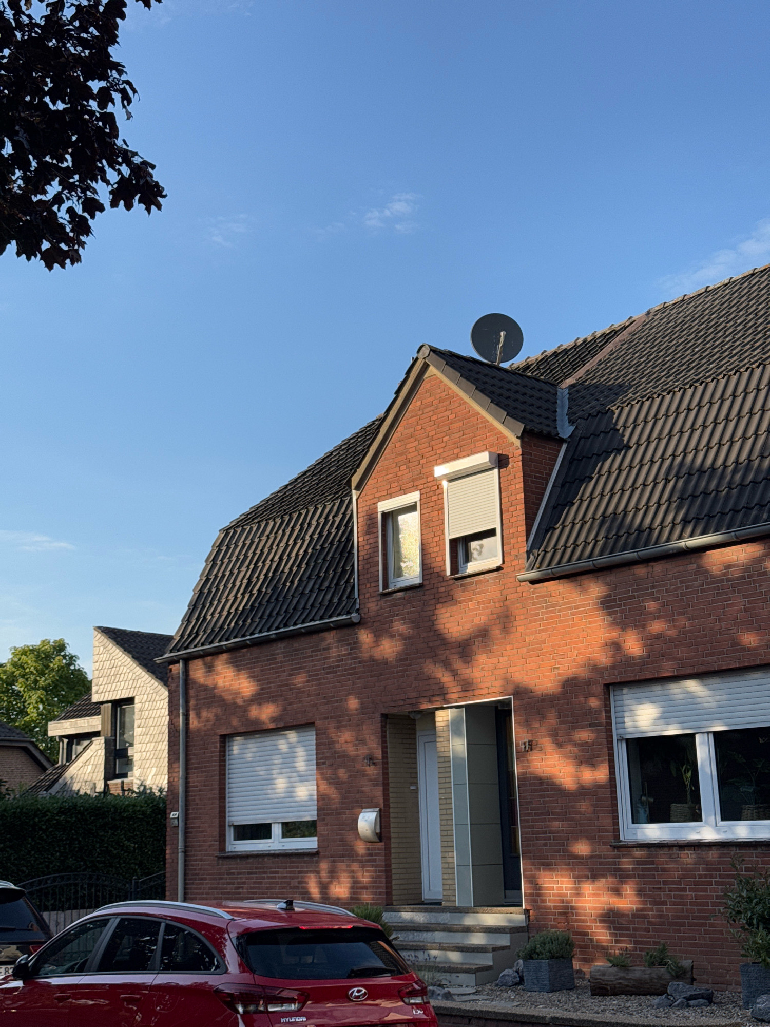 The image shows the exterior of a residential house in Cleves, Germany. The house is constructed of red brick with a dark-tiled roof. A satellite dish is mounted on the roof. The windows have roller shutters, some of which are down. There is a small set of stairs leading to the front door, which is partially open, revealing a glimpse of the interior. Shadows from trees are cast across the brick facade. In the foreground, a red car is parked. The sky is clear and blue.