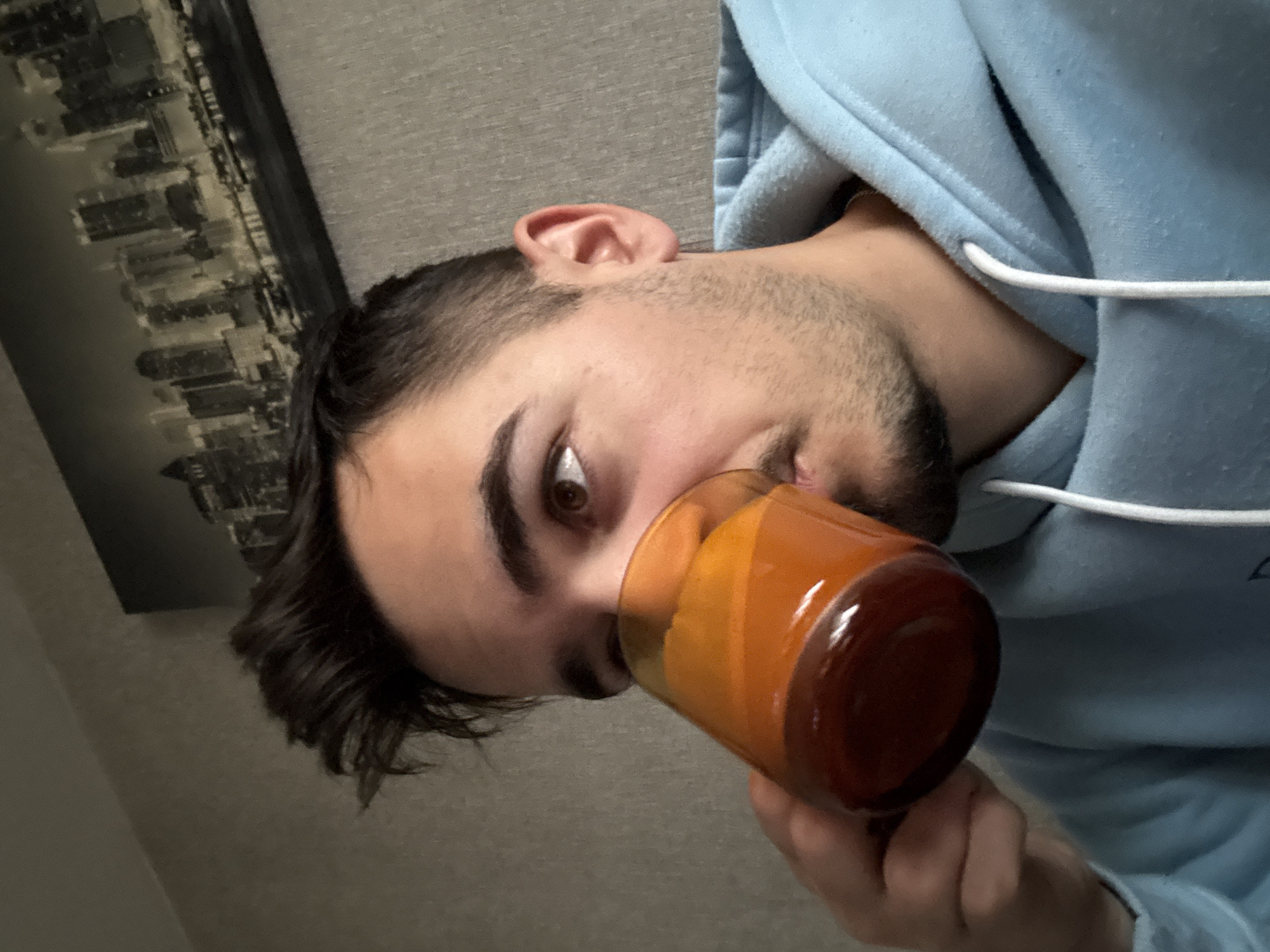 A man is drinking from a brown and orange glass. He is wearing a light blue hoodie. A picture of a city skyline hangs on the wall behind him. It is not possible to determine the location or city of Sofia, Bulgaria.
