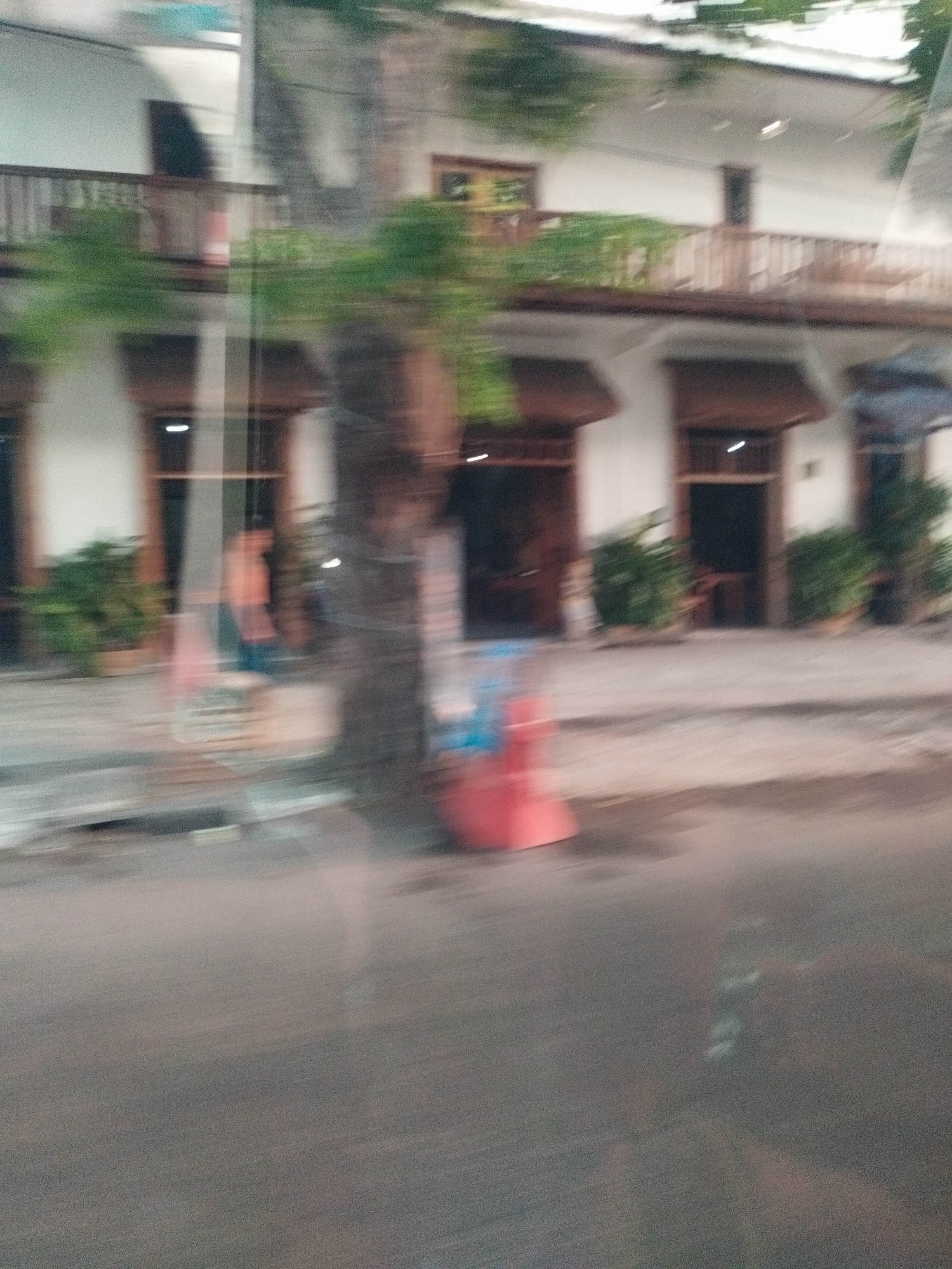 The image shows a blurred view of a building with a balcony and several doorways. Plants are visible on the ground floor. A tree trunk stands prominently in the foreground. The scene appears to be a street in Surabaya, Indonesia, possibly at night or during low light conditions, given the artificial lighting visible within the doorways. The overall impression is one of movement, with the blur suggesting the photograph was taken from a moving vehicle. There are no discernible interactions or specific actions taking place.