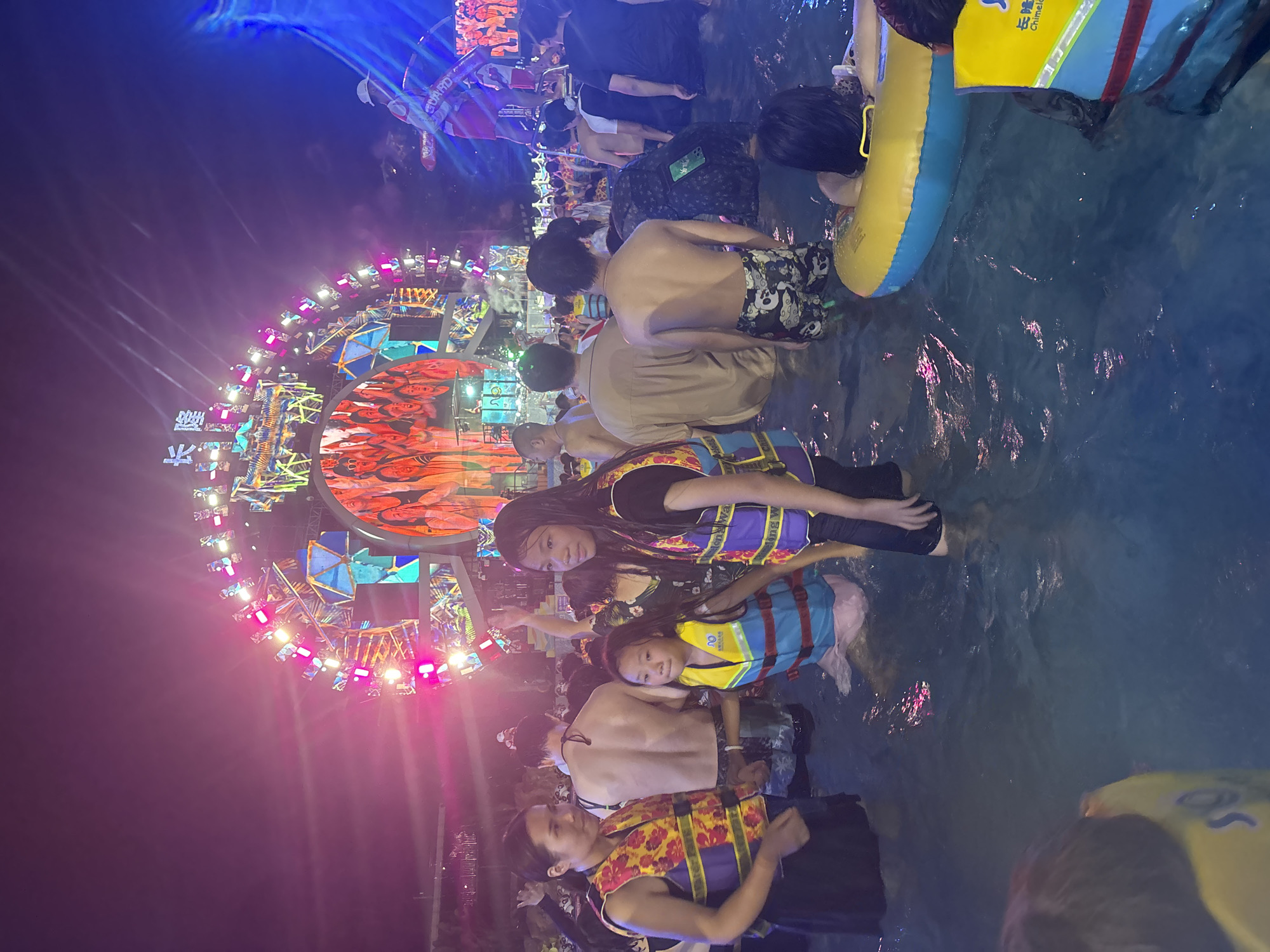 In the city of Panyu District, China, a crowded water park is depicted at night. A large, brightly lit stage structure dominates the background, featuring a circular screen displaying images of people. The stage is adorned with numerous colorful lights and geometric shapes. In the foreground, numerous individuals are standing in the water, many wearing life vests of various colors, including yellow, blue, and pink. Some people are in groups, engaged in conversation, while others appear to be observing the stage. The water is illuminated by the vibrant lights from the stage, creating reflections on its surface. The atmosphere suggests a lively event, possibly a concert or festival, taking place at the water park.