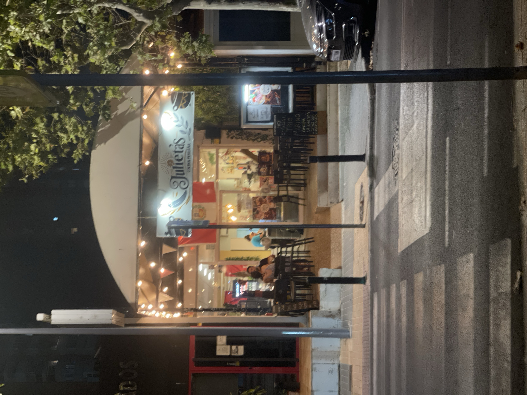The image shows a pedestrian crossing in front of a restaurant named "Julieta's Cocina Peruana" in Benidorm, Spain. The restaurant has outdoor seating with tables and chairs, and is decorated with string lights and pennant banners. Inside, the restaurant is brightly lit, and several patrons are visible. A dark-colored car is parked on the right side of the image. The scene is at night, with the street and sidewalk illuminated by streetlights and the restaurant's lighting. A tree with green foliage is visible on the right side of the image, casting shadows.