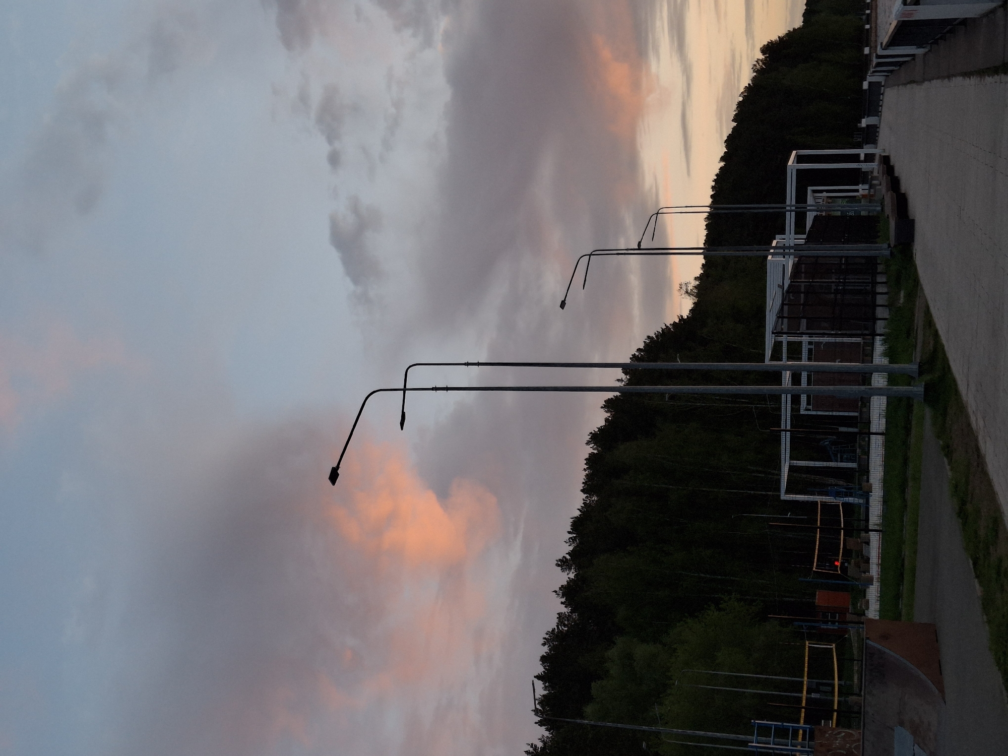 The image shows a street in the city of Chaikovsky, Russia, at dusk. A row of street lamps line the road. A forest borders the street on one side, and a pedestrian walkway is on the other. A building with a white frame is adjacent to the walkway. The sky is filled with pink and gray clouds.