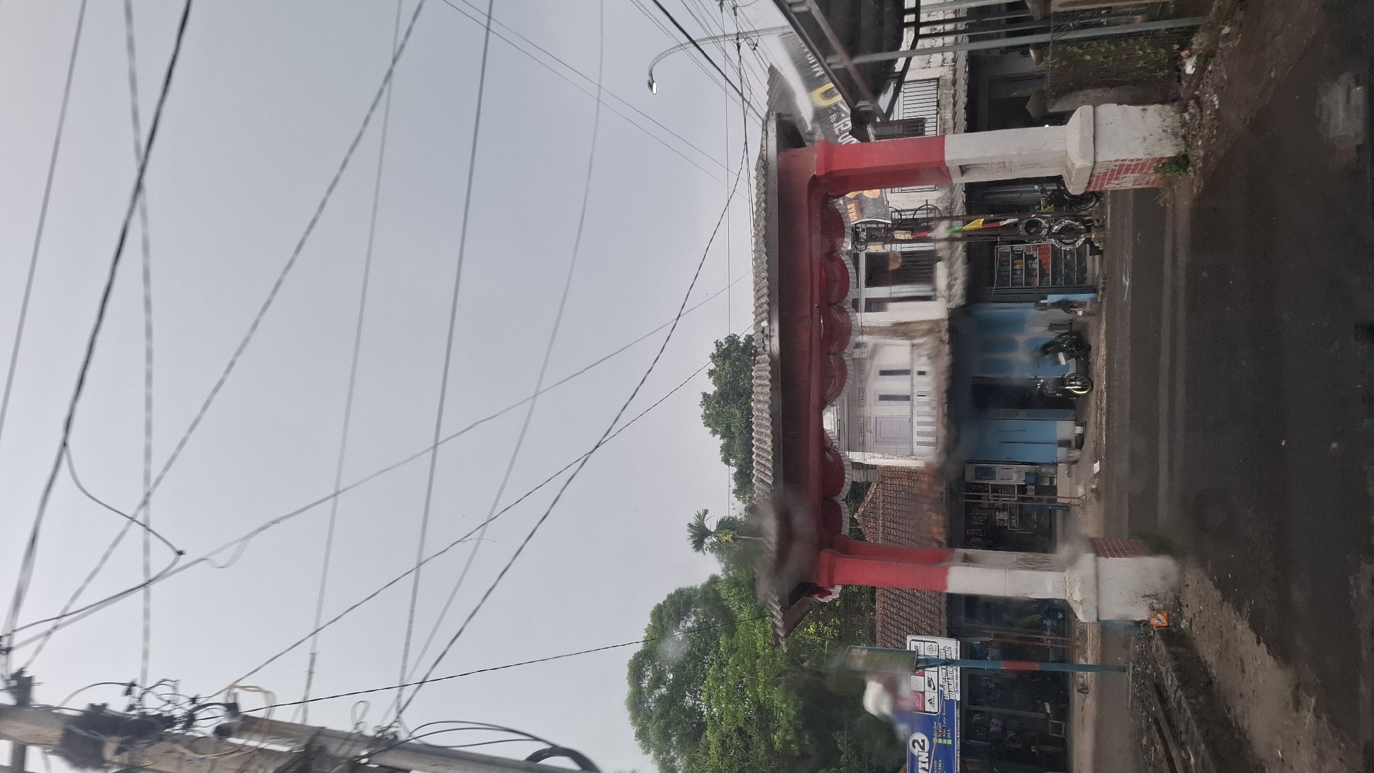 A street-level, medium shot captures a red and white arched gateway decorated with red fabric draped across its top. The gateway stands on a wet asphalt road in Sukasari, Indonesia, under an overcast sky. To the left of the gateway, a utility pole is laden with numerous electrical wires that crisscross the frame. Behind the gateway, a two-story building features a light blue facade on the ground floor with several shops or entrances. Two motorcycles are parked on the pavement in front of the building, one white and one black. To the right of the gateway, another building structure is visible, partially obscured by the gateway. Lush green trees are present in the background, beyond the buildings. The overall scene suggests a typical street view in a residential or commercial area.