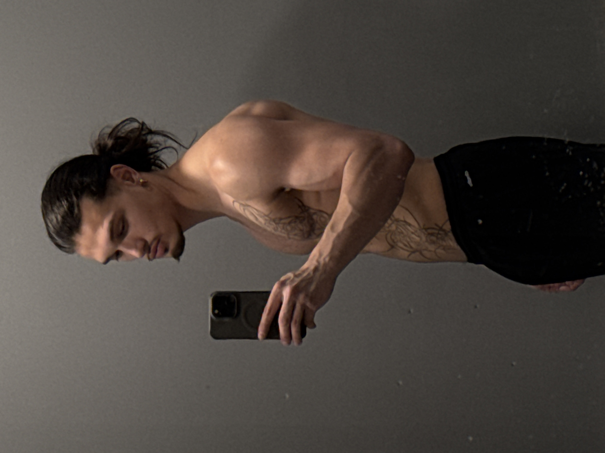 A shirtless man with dark hair pulled back in a bun and a goatee is standing in front of a gray wall, taking a mirror selfie with a dark-colored smartphone. He has tattoos on his torso and is wearing black pants. The setting appears to be a gym or a similar indoor location with plain walls. There are no discernible landmarks or environmental features to indicate a specific location within Sofia, Bulgaria.