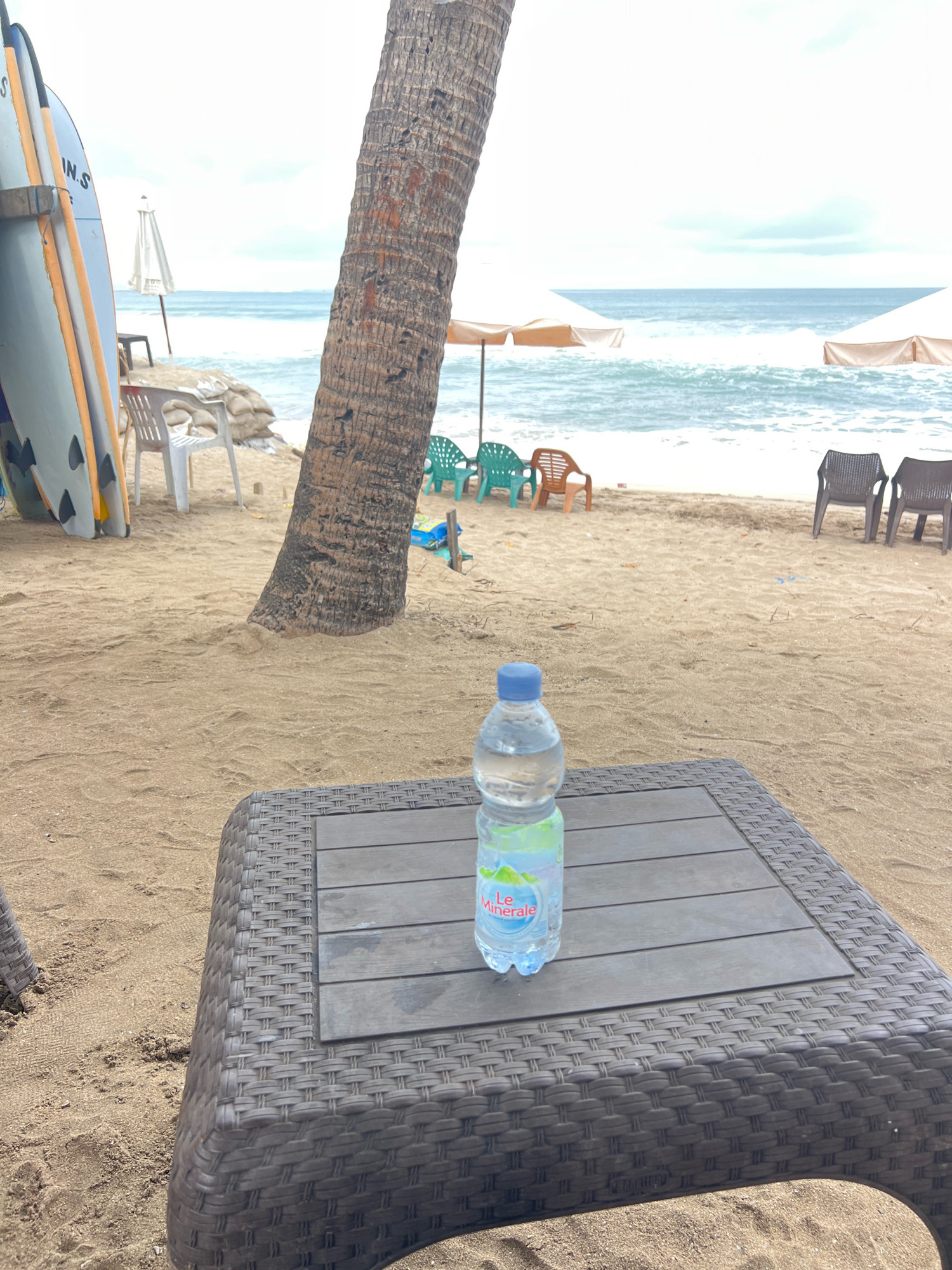 A bottle of "Le Minerale" water sits on a dark brown woven plastic table on a sandy beach in Kuta, Indonesia. In the background, the ocean waves crash onto the shore under a cloudy sky. A palm tree trunk is visible to the left of the bottle, and several beach chairs and surfboards are scattered across the sand. Two large beige beach umbrellas are partially visible on the right side of the frame.