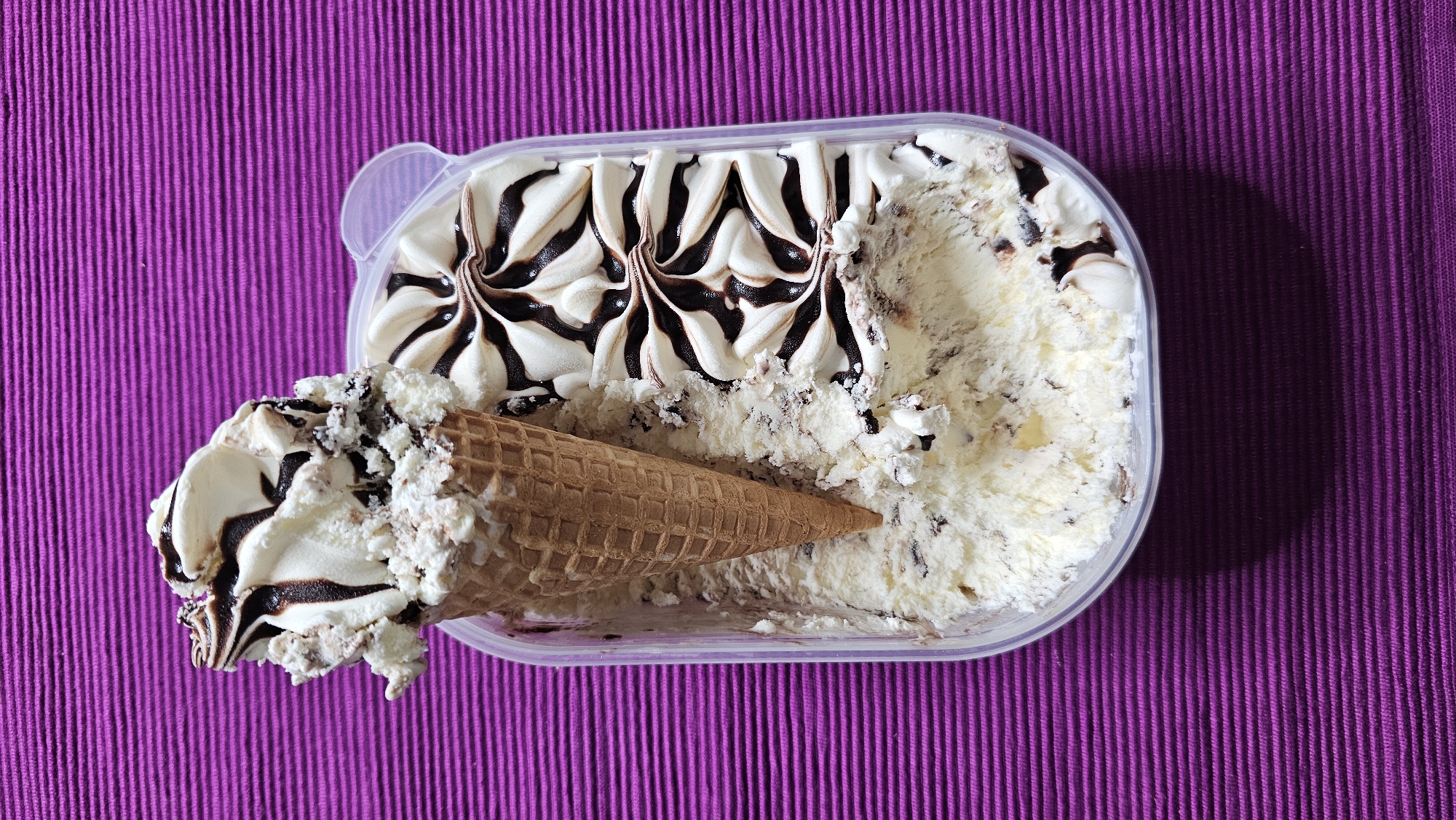 A high-angle, close-up forensic photograph depicts a clear plastic rectangular container of ice cream, positioned on a textured, horizontally ridged purple fabric surface, in the city of Boussu-lez-Walcourt, Belgium. The container holds approximately one gallon of vanilla-based ice cream with dark chocolate chunks or ripples. The upper right portion of the ice cream within the container features decorative swirled peaks of white ice cream liberally drizzled with dark chocolate syrup. An inverted waffle ice cream cone, filled with a scoop of the same vanilla and chocolate chunk ice cream, is centrally embedded into the container's contents. This cone-filled scoop is heavily topped with additional dark chocolate syrup and scattered white crumbs or flakes. A distinct shadow is cast by the container onto the purple surface, originating from the upper left. No individuals or direct interactions are visible.
