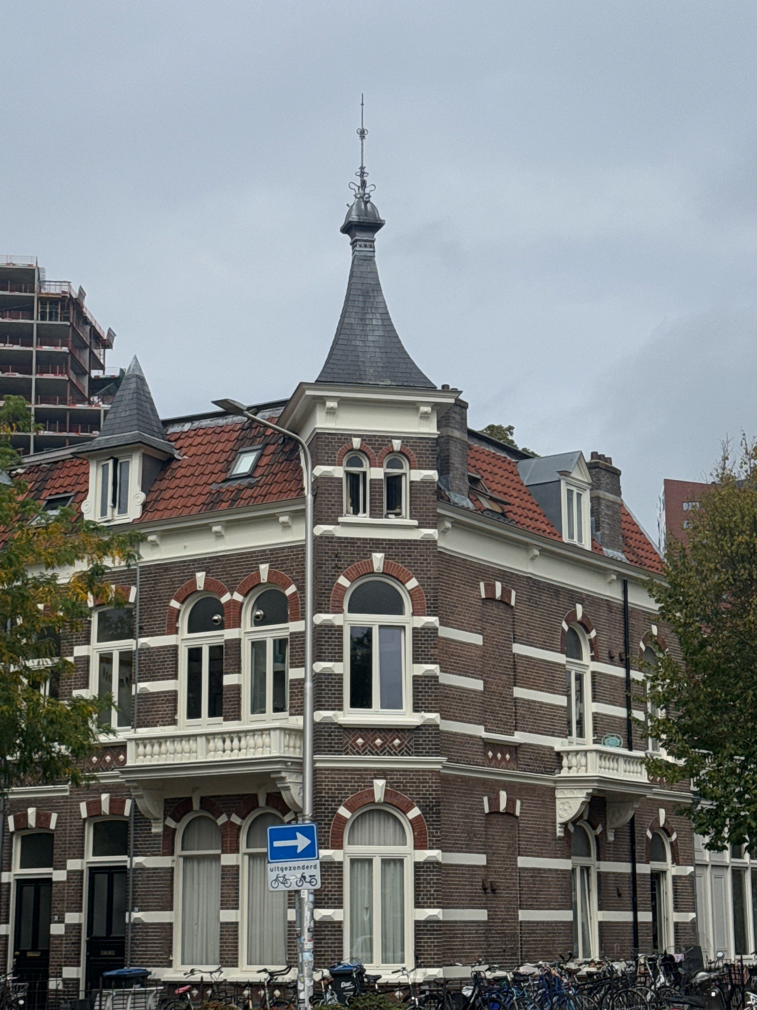The image shows a corner view of a multi-story brick building with red tile roofs and white trim, located in the city of Nijmegen, Netherlands. The building features arched windows, balconies, and decorative brickwork. A tall, conical turret with a decorative spire tops one corner of the building. To the left of the building, a partially constructed tall building with exposed concrete framework is visible against an overcast sky. In the foreground, a dense collection of parked bicycles is visible along the street. A street sign indicating a rightward arrow and symbols for bicycles is mounted on a pole. Trees with autumnal foliage frame the right side of the building. The overall atmosphere is urban and overcast.