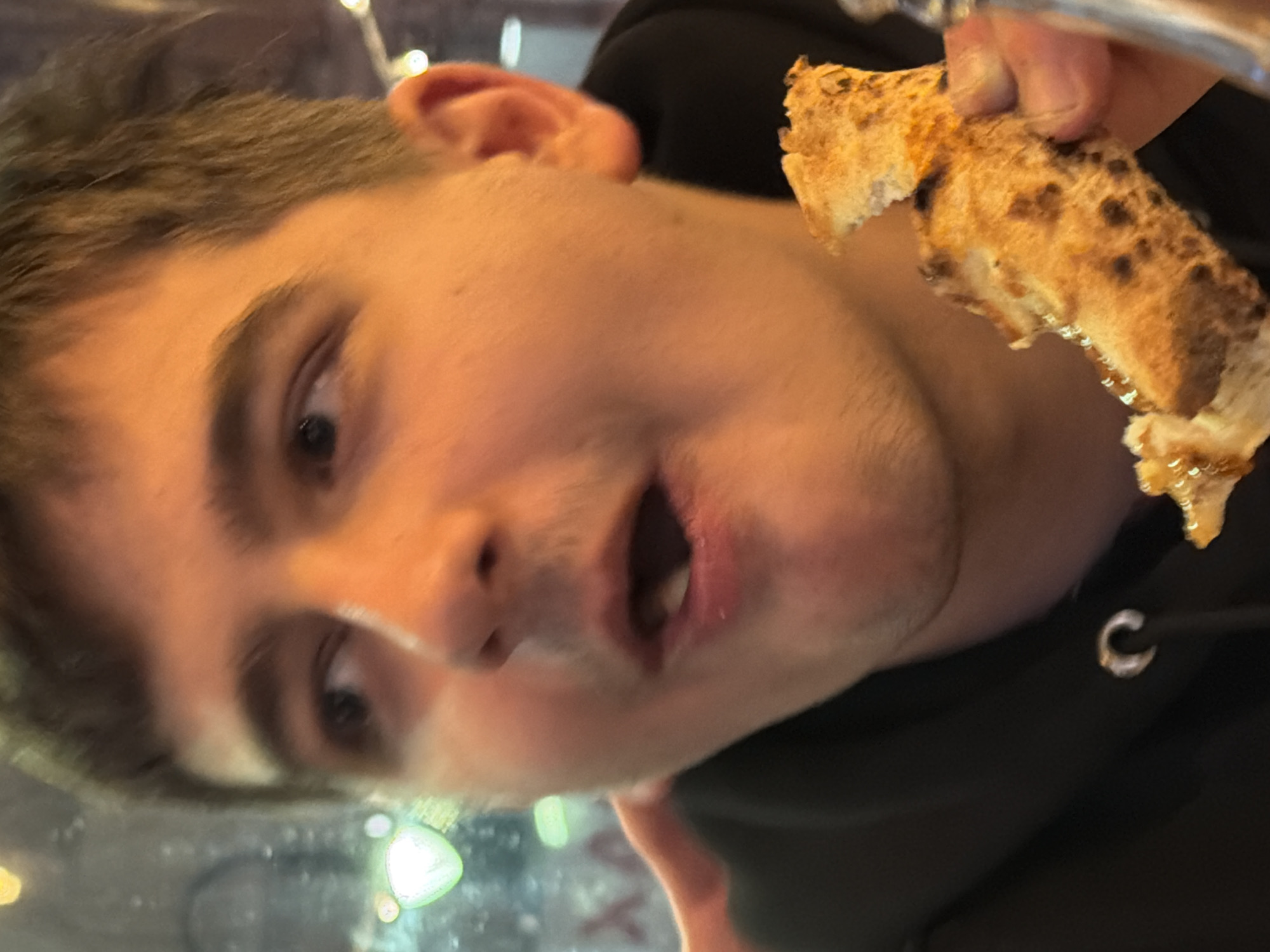 A close-up, candid depiction of a young adult male in a casual dining establishment in Sofia, Bulgaria. The subject, centered in the frame, possesses short, dark brown hair and a light amount of facial stubble. His dark eyes are directed towards the upper left, and his mouth is open, revealing teeth, consistent with speech or the act of eating. He is wearing a dark, likely black, hooded garment, with a silver-colored eyelet visible on the neckline. His right hand holds a partially consumed slice of pizza, identifiable by its charred crust and visible toppings, positioned near his open mouth. The background is out of focus, showing multiple bokeh light sources from string lights and other ambient fixtures, indicating an evening setting. Faint, blurred red Cyrillic characters, specifically "XA", are visible through a window or reflection on the left side of the background, consistent with a commercial establishment in a Bulgarian city.