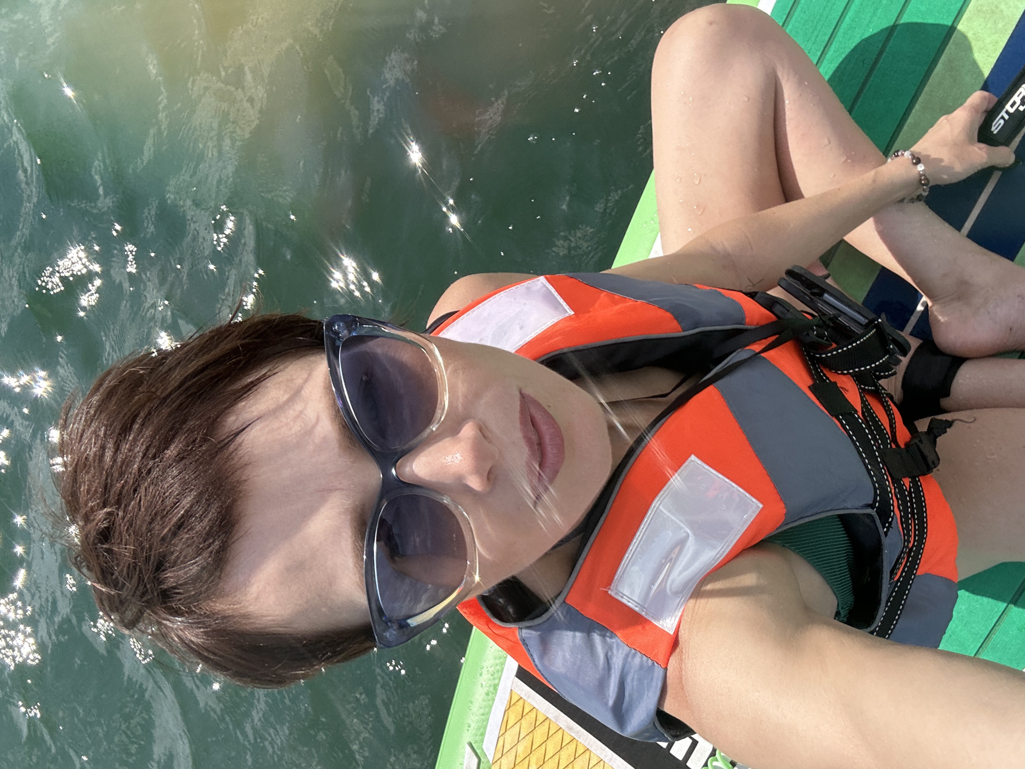 A woman is taking a selfie while sitting on a paddleboard in the water. She is wearing sunglasses and an orange and gray life vest over a green swimsuit. The water is green and reflects the sunlight, creating a shimmering effect. The paddleboard has green and white stripes. The woman has short brown hair and is looking at the camera with a slight smile. The image appears to be taken on a sunny day. The person is sitting with their legs crossed. A black paddle handle is visible in the upper right corner of the image. Text visible on the paddle handle reads "STORM".