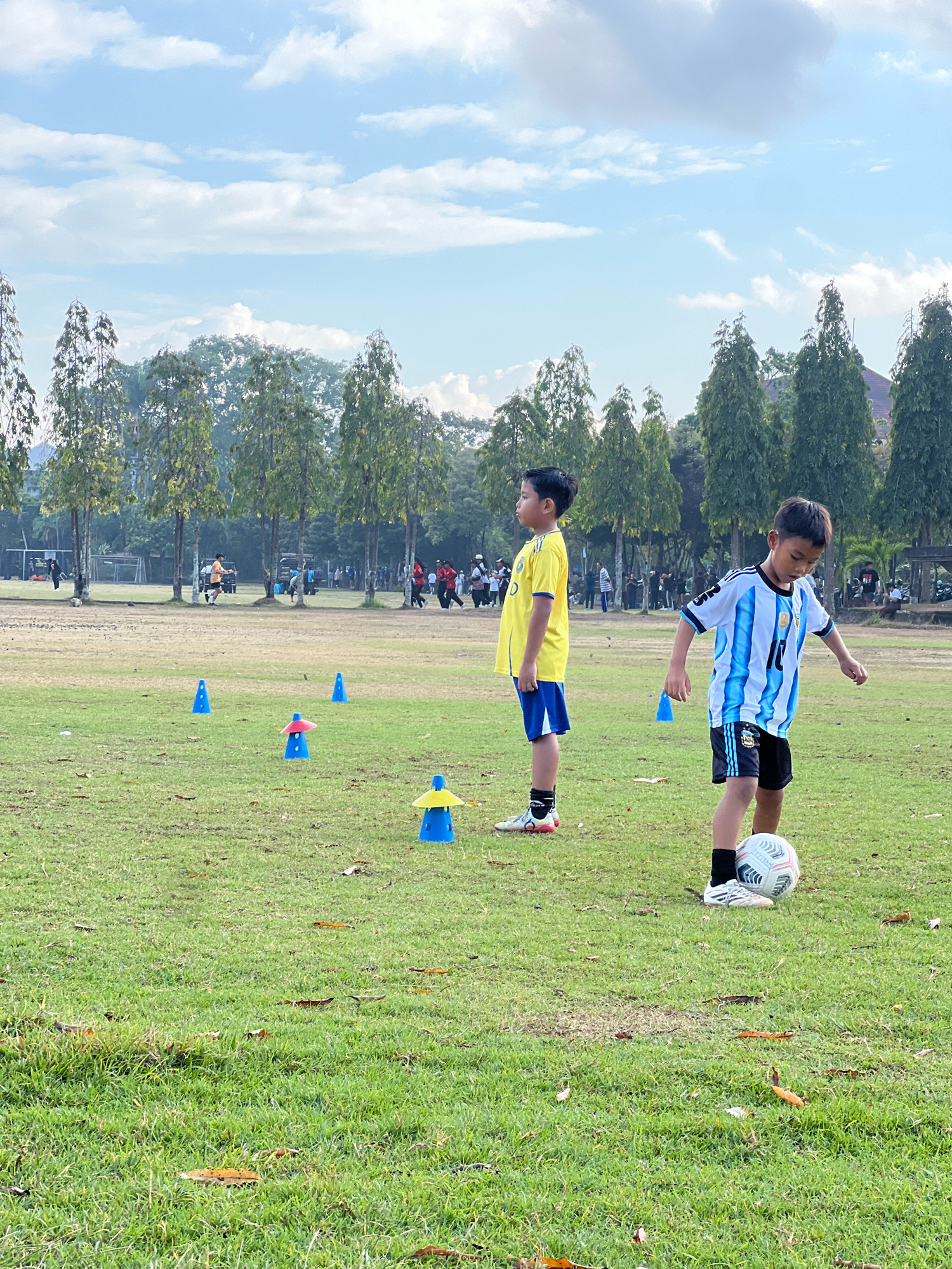 The image shows two young boys on a grassy field, likely at a sports training facility in Denpasar, Indonesia. The boy in the foreground is wearing an Argentina national football team jersey and shorts and is playing with a soccer ball. The other boy, standing to his left, is wearing a yellow and blue soccer jersey and shorts. There are several blue and yellow cone markers on the field, suggesting a training drill. In the background, a line of trees borders the field, and further back, there are glimpses of buildings and other people. The sky is partly cloudy with a light blue hue.