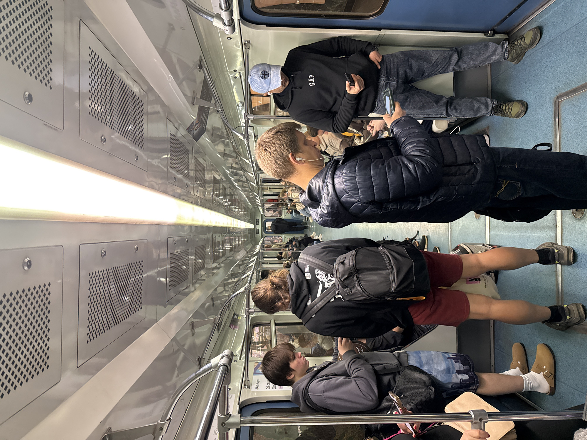 Inside a Moscow Metro train, Russia. Passengers are standing and sitting along both sides of the carriage. Many are using their phones. The train is illuminated by a long strip light along the ceiling. Seating is blue. Walls are a light grey.