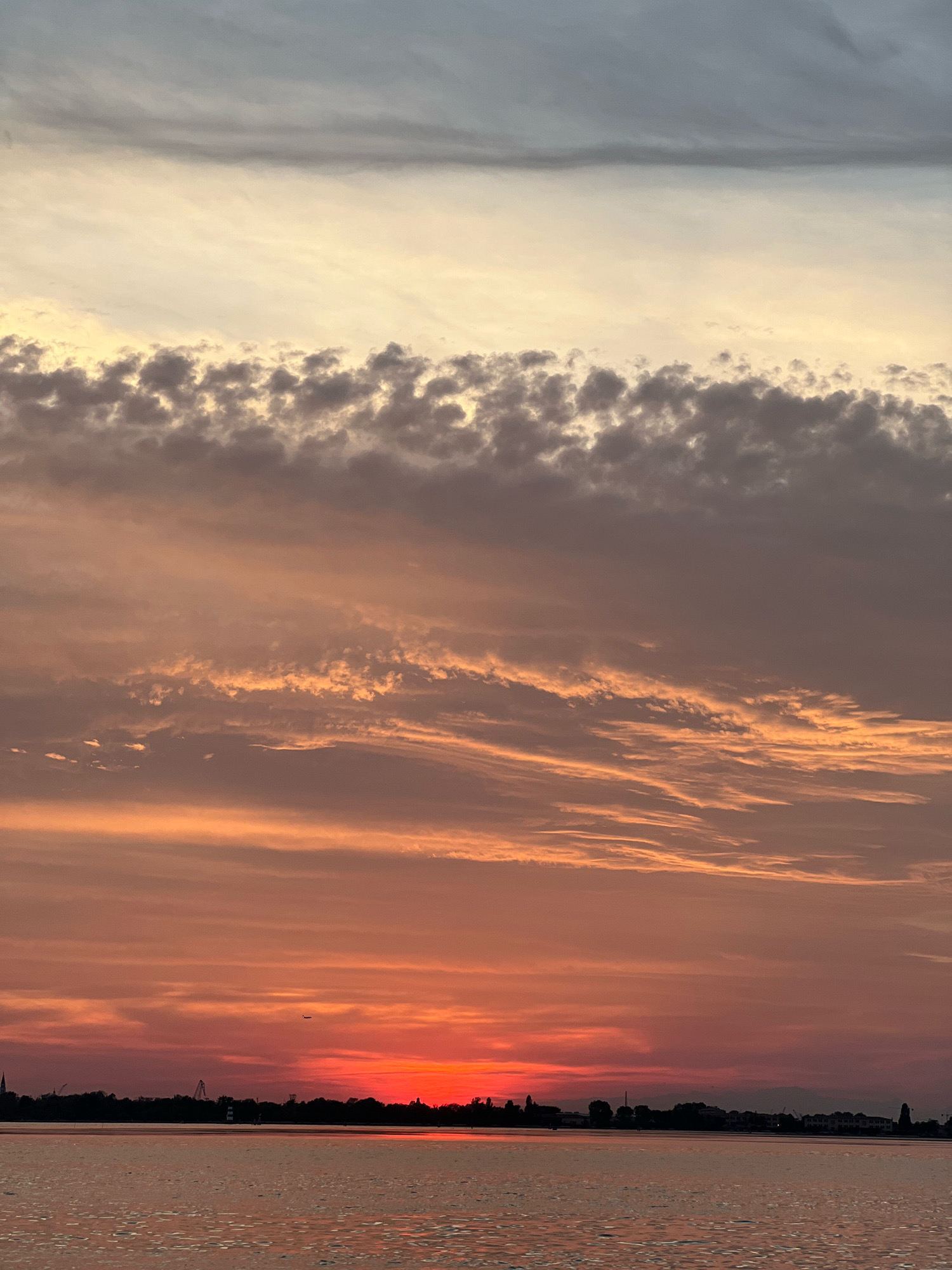 This image shows a sunset over the Venetian Lagoon, as viewed from Punta Sabbioni, Italy. The sky is filled with dramatic clouds in shades of orange, pink, and gray, with a bright red glow near the horizon where the sun is setting. The water in the foreground is calm and reflects the colors of the sky. A silhouetted shoreline with buildings, trees, and a lighthouse is visible in the distance. A small airplane is flying in the sky.