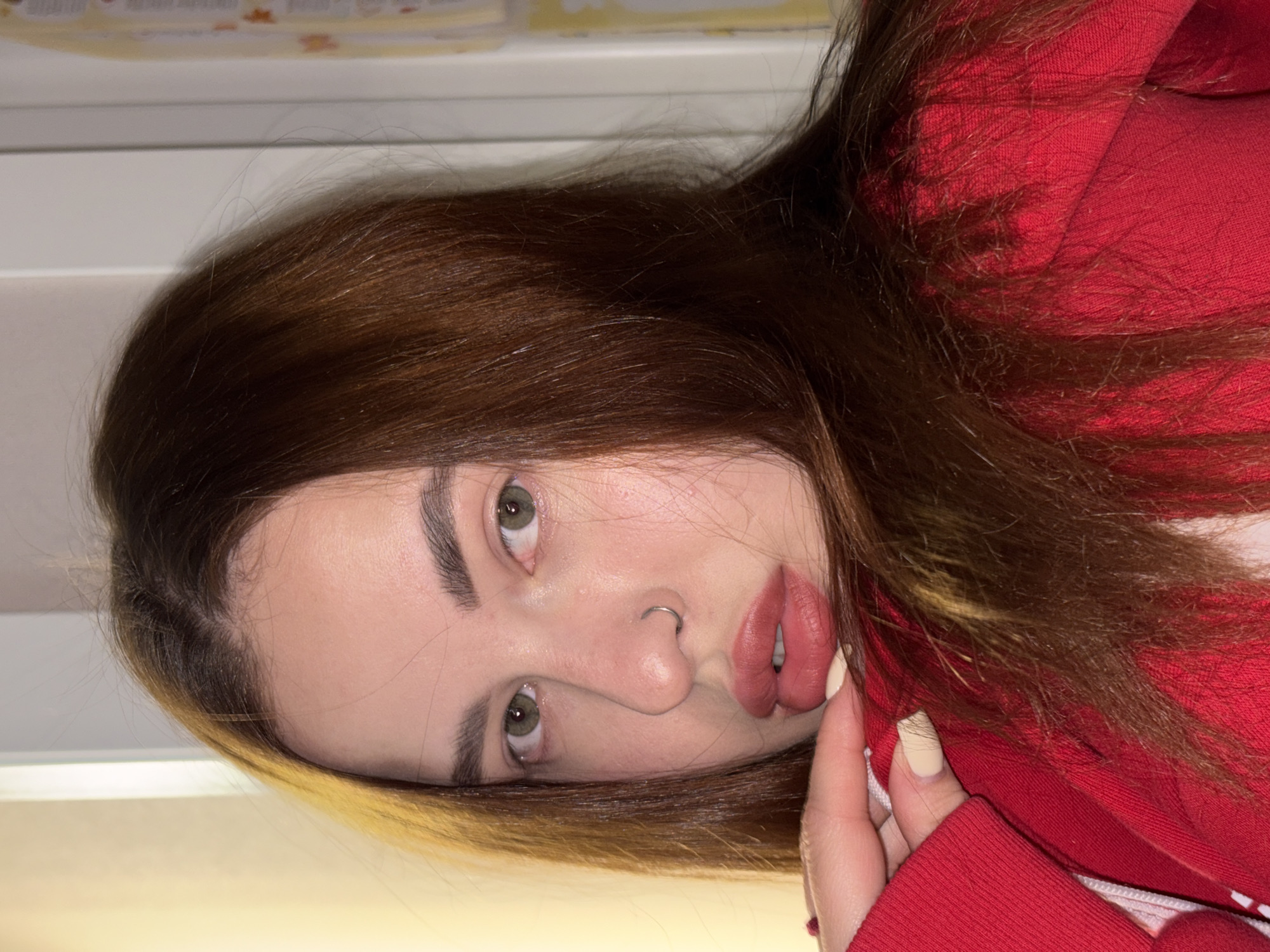 The image is a close-up portrait of a young person, likely female, rotated ninety degrees to the left. The subject has long, dark brown hair with some lighter, possibly dyed, streaks visible towards the front. Their green eyes are directed towards the viewer, and they have light skin and prominent eyebrows. A silver hoop nose ring is visible on their left nostril. Their lips are full and painted with a muted pink or mauve lipstick.
The person is wearing a red hooded sweatshirt or jacket, the textured fabric of which is prominent. One hand, with light-colored (possibly cream or pale yellow) nail polish, is holding a portion of the red garment near their chin. Loose strands of hair are visible on the red fabric of the hoodie.
The background is uniformly light-colored and out of focus, suggesting an indoor setting. At the top of the frame, some faint, blurry rectangular shapes are visible, possibly indicating a shelf or architectural feature, but no specific details or discernible text can be made out. The lighting appears soft and even, highlighting the subject's features. The overall expression is neutral and direct, characteristic of a posed portrait. The context provided indicates the location is Krasnodar, Russia.