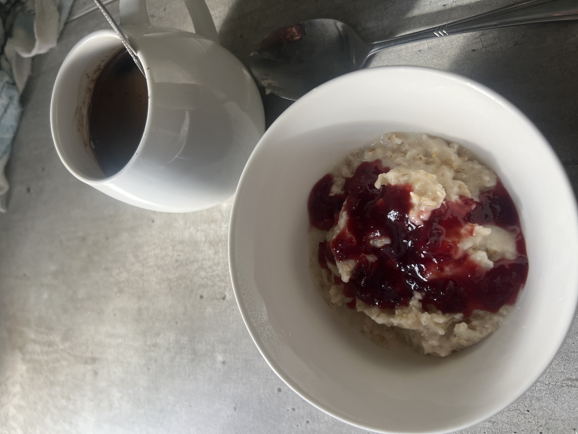 A bowl of oatmeal topped with jam and a dollop of cream sits on a grey, textured countertop. Next to the bowl is a white mug filled with dark coffee, and a spoon rests beside the bowl. The scene is illuminated by natural light, casting shadows. The image does not provide any information about the city of Volgograd, Russia, or any specific location.