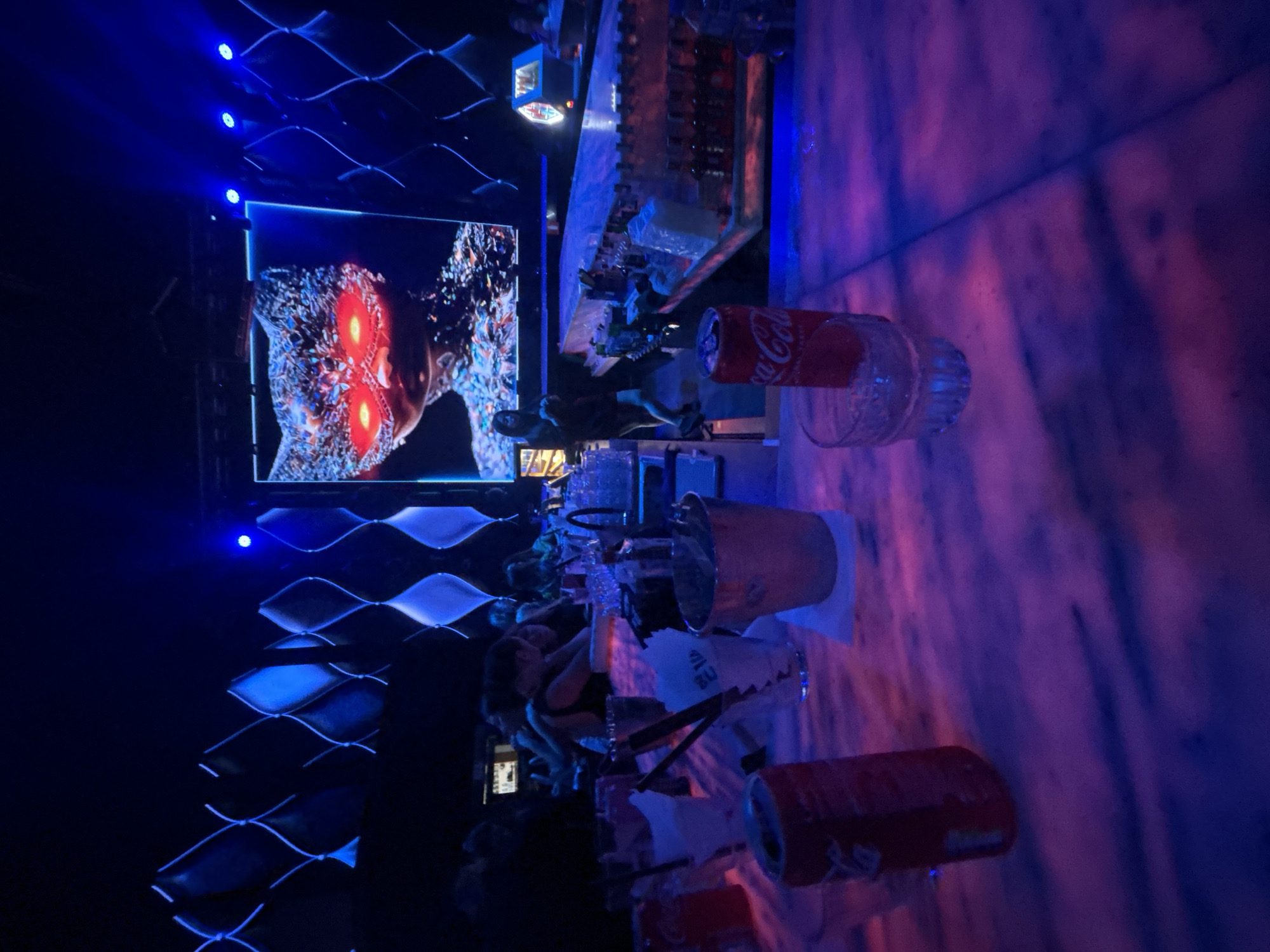 The image depicts a dimly lit nightclub or bar environment, bathed in blue and pink neon lighting. In the foreground, a marble-topped bar displays several drink containers, including two cans of Coca-Cola and an ice bucket. A large screen in the background features a digital image of a person wearing a bejeweled cat mask with glowing red eyes. Several patrons are visible in the background, some seated at the bar and others standing. The overall atmosphere suggests a vibrant nightlife scene. The image location is not specified in the original prompt nor can it be deduced from the visual elements.